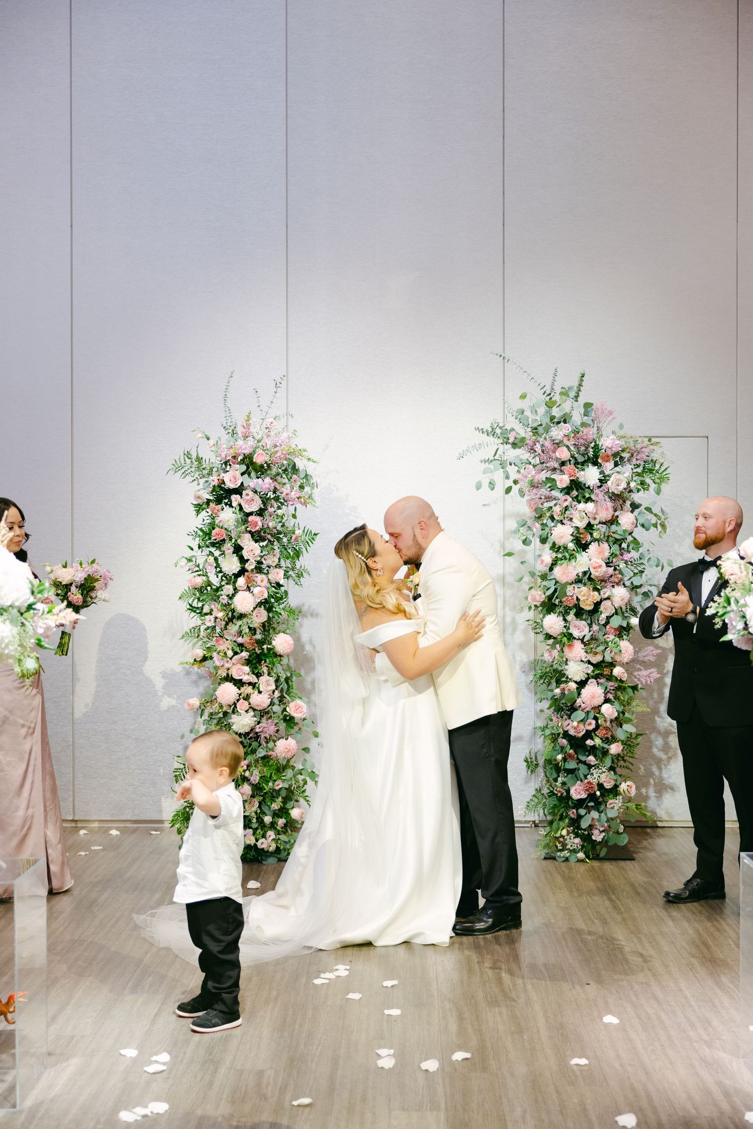 Wedding celebration with couple kissing under floral arch as guests and child look on.