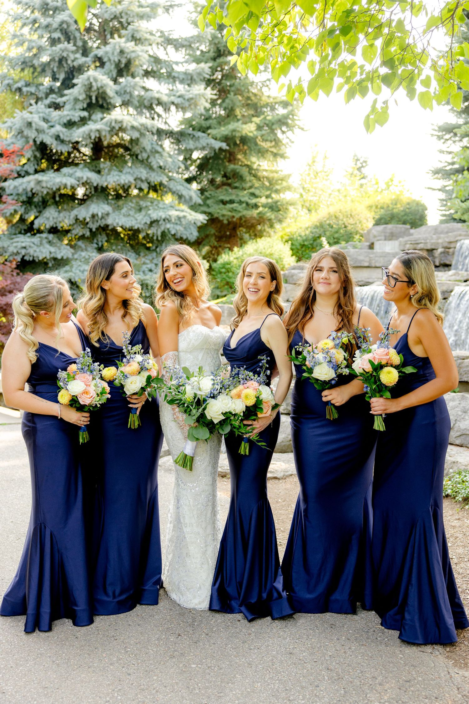 A bridal party in navy blue dresses and a white wedding gown poses together while holding floral bouquets outdoors.