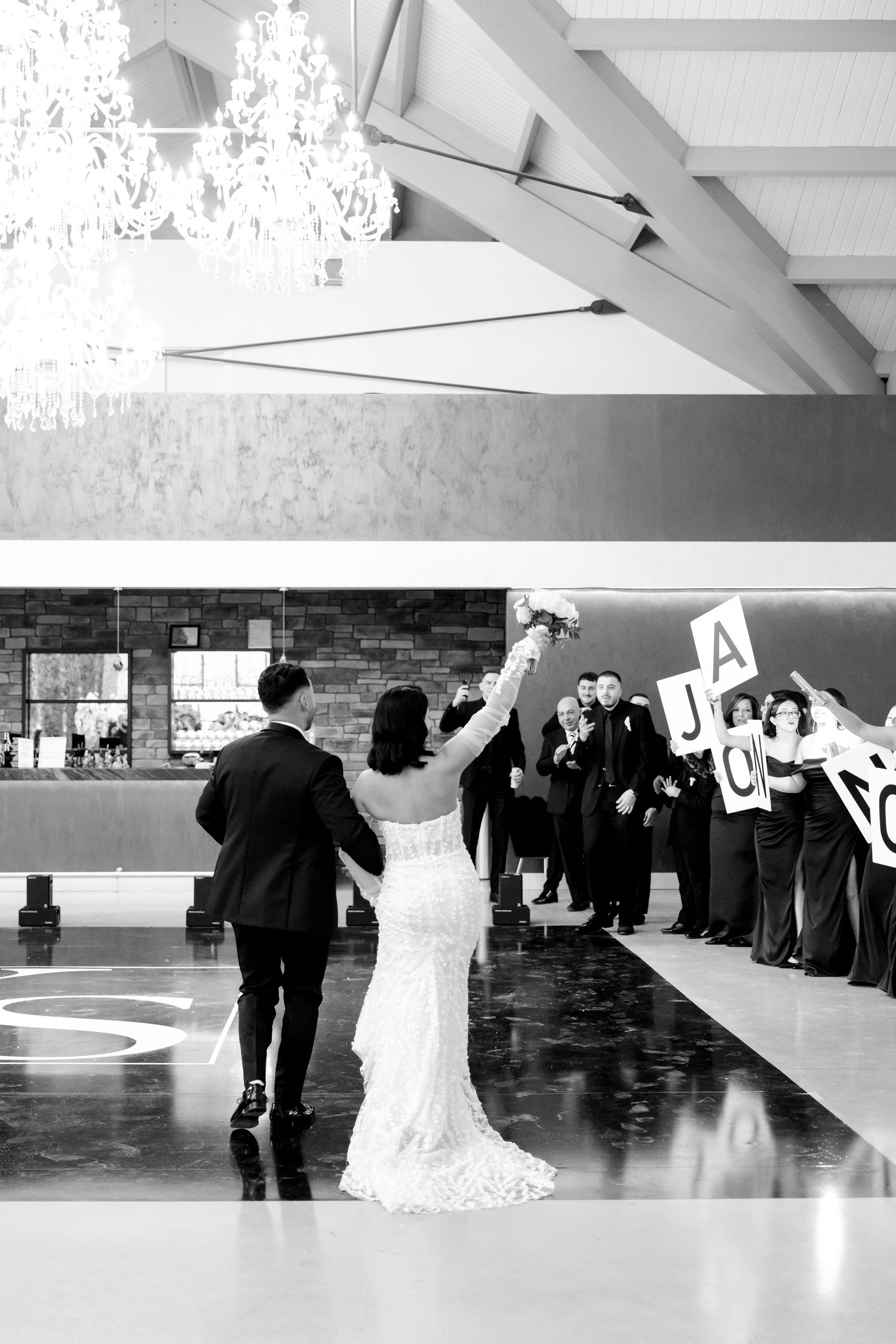 Black and white photo of newlyweds walking past guests holding letter signs at their reception.