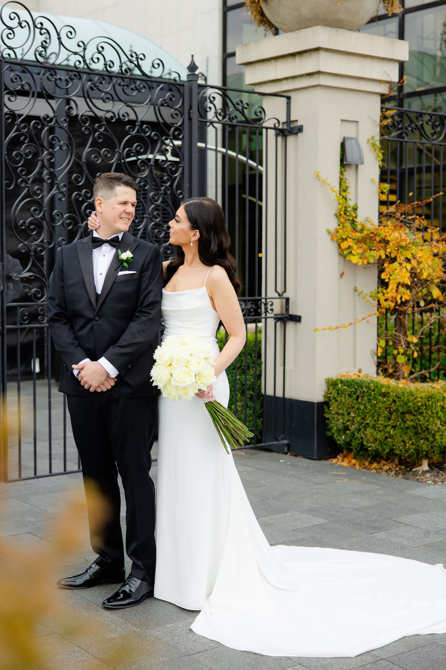 Wedding couple stands before ornate gates with yellow autumn foliage in background.
