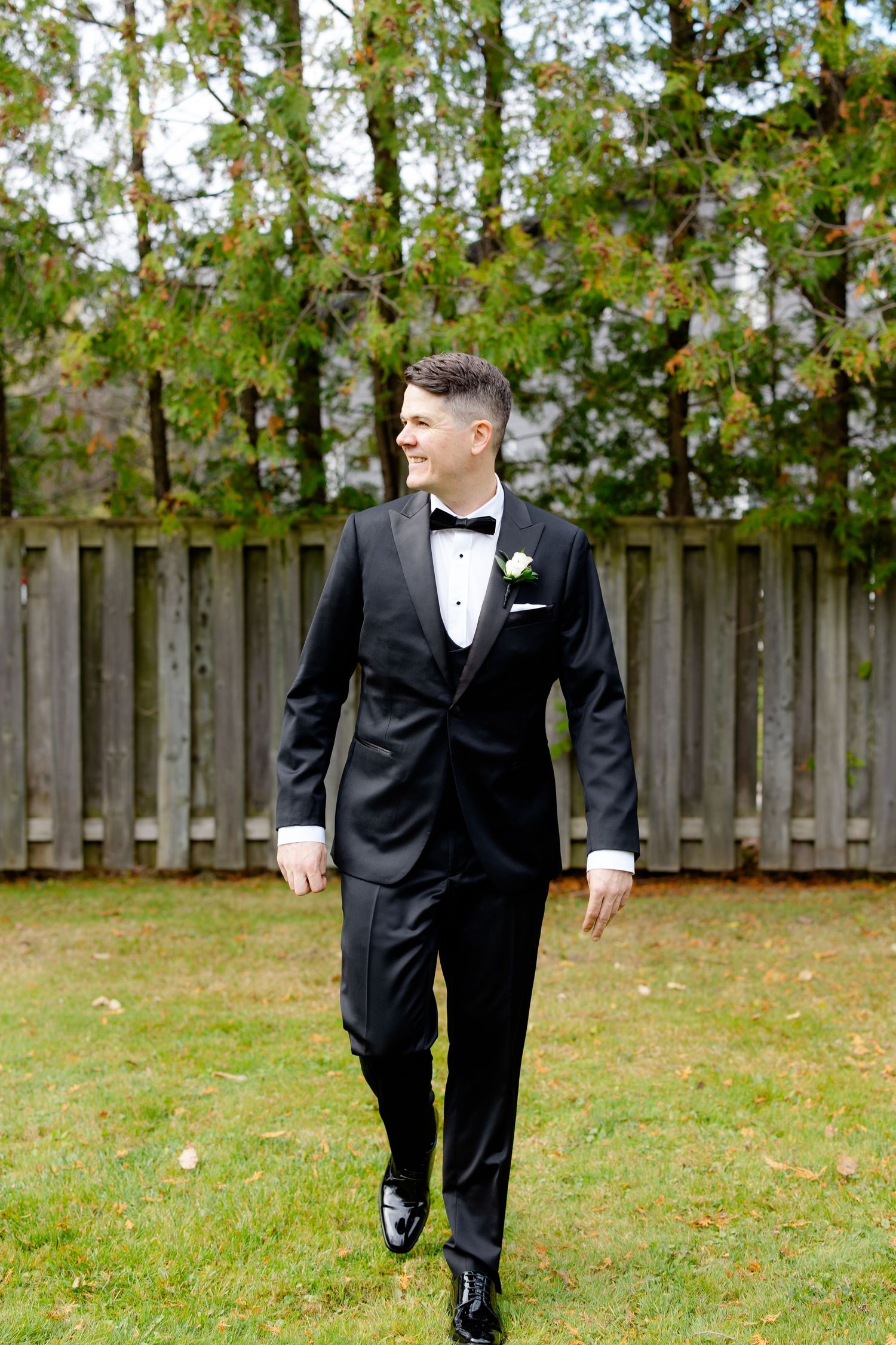 A groom in a classic black tuxedo walks across a lawn with a wooden fence and trees in the background.