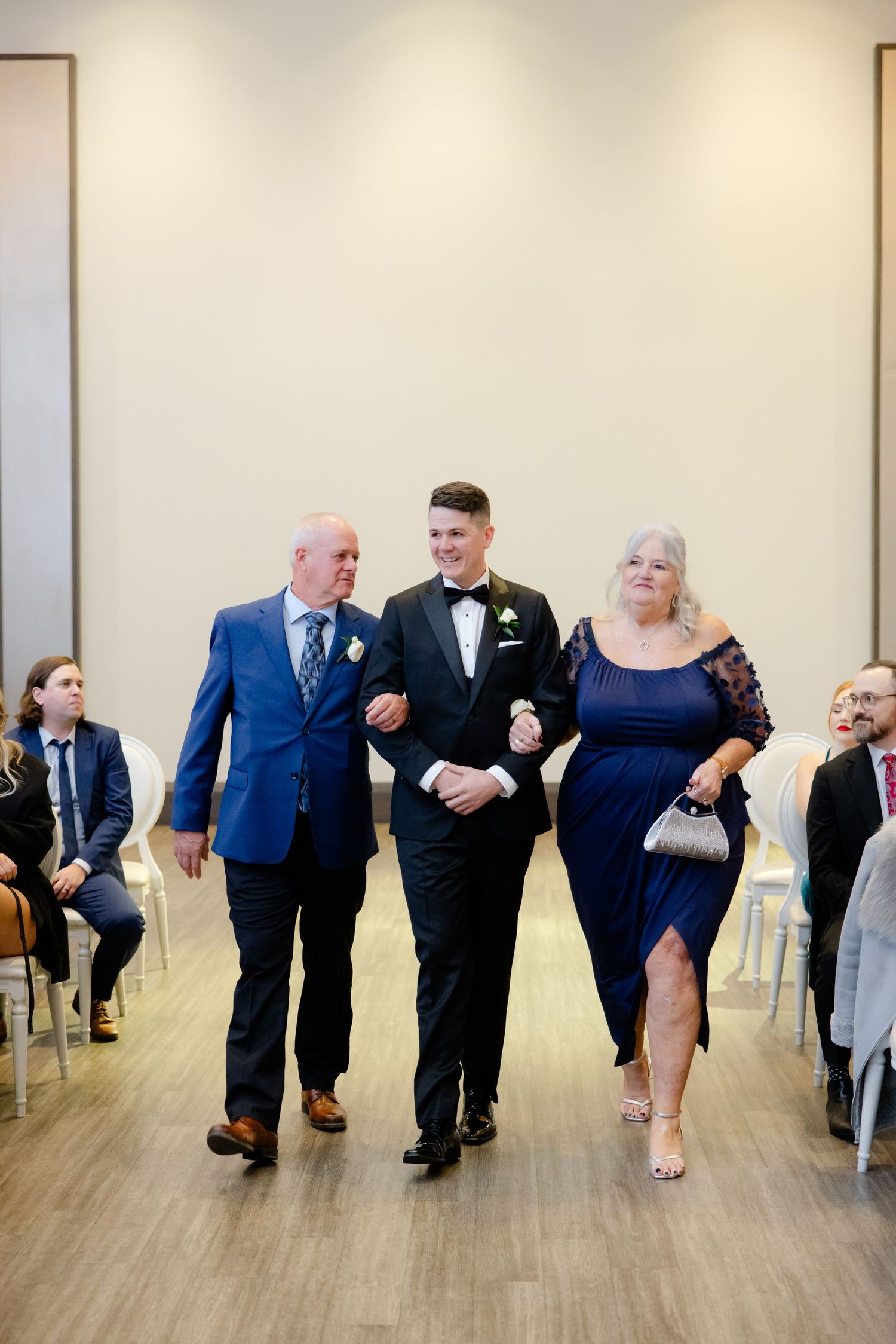 Wedding party members walk down the aisle in coordinated navy and black formal attire at an elegant indoor ceremony.