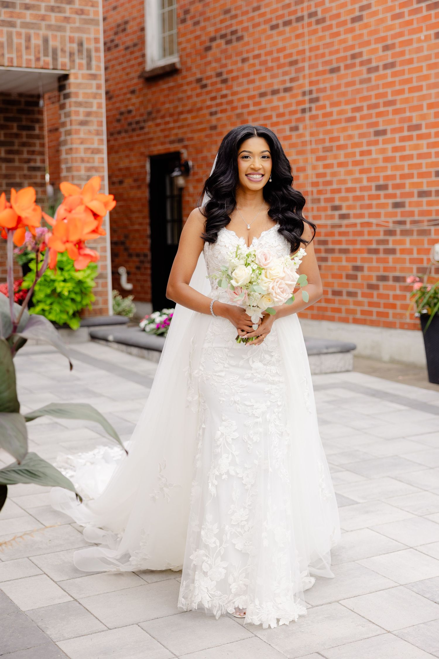 Beautiful bride in lace wedding dress holds white bouquet against brick building exterior.