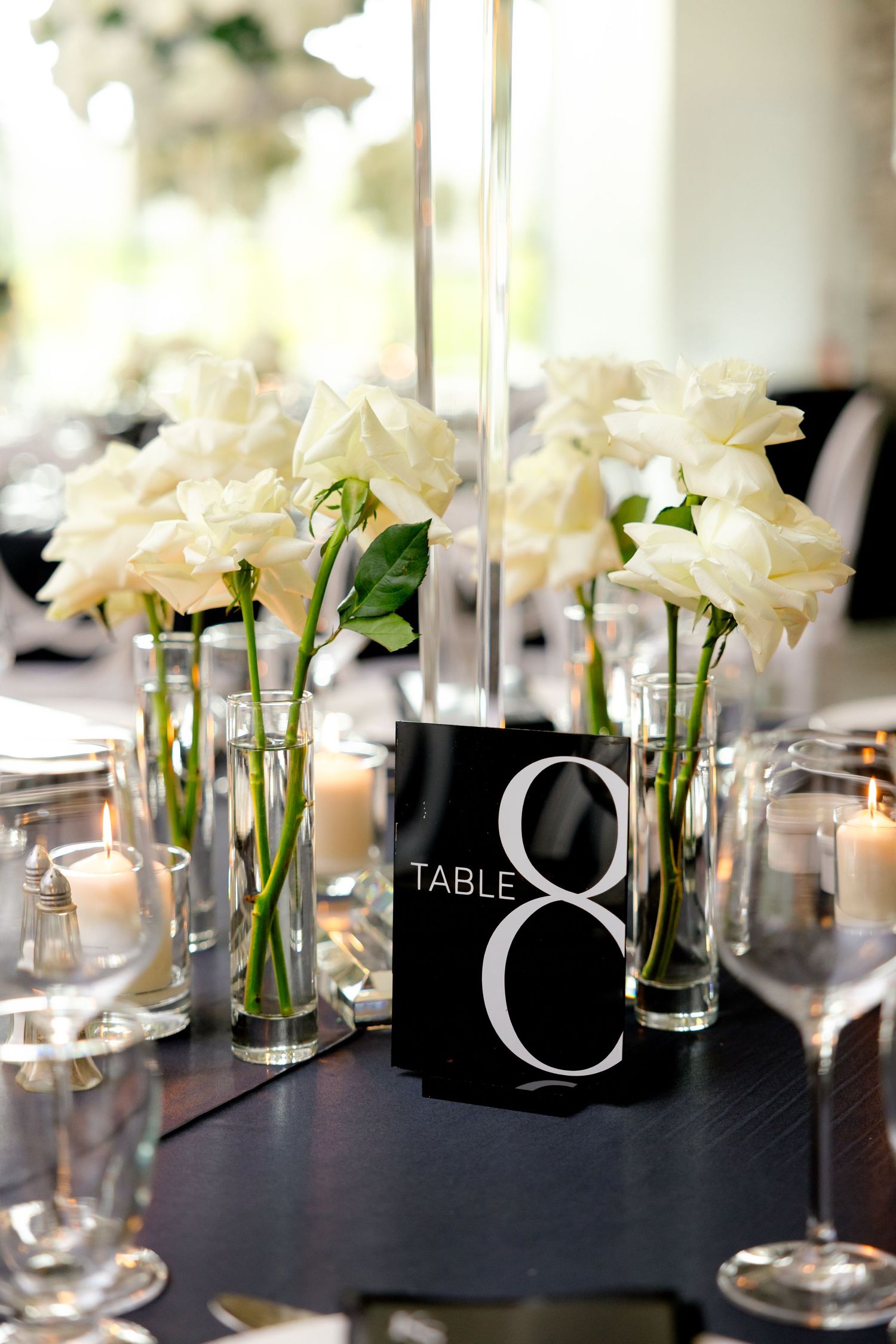 Modern table setting with white roses in glass vases and a black table number card.