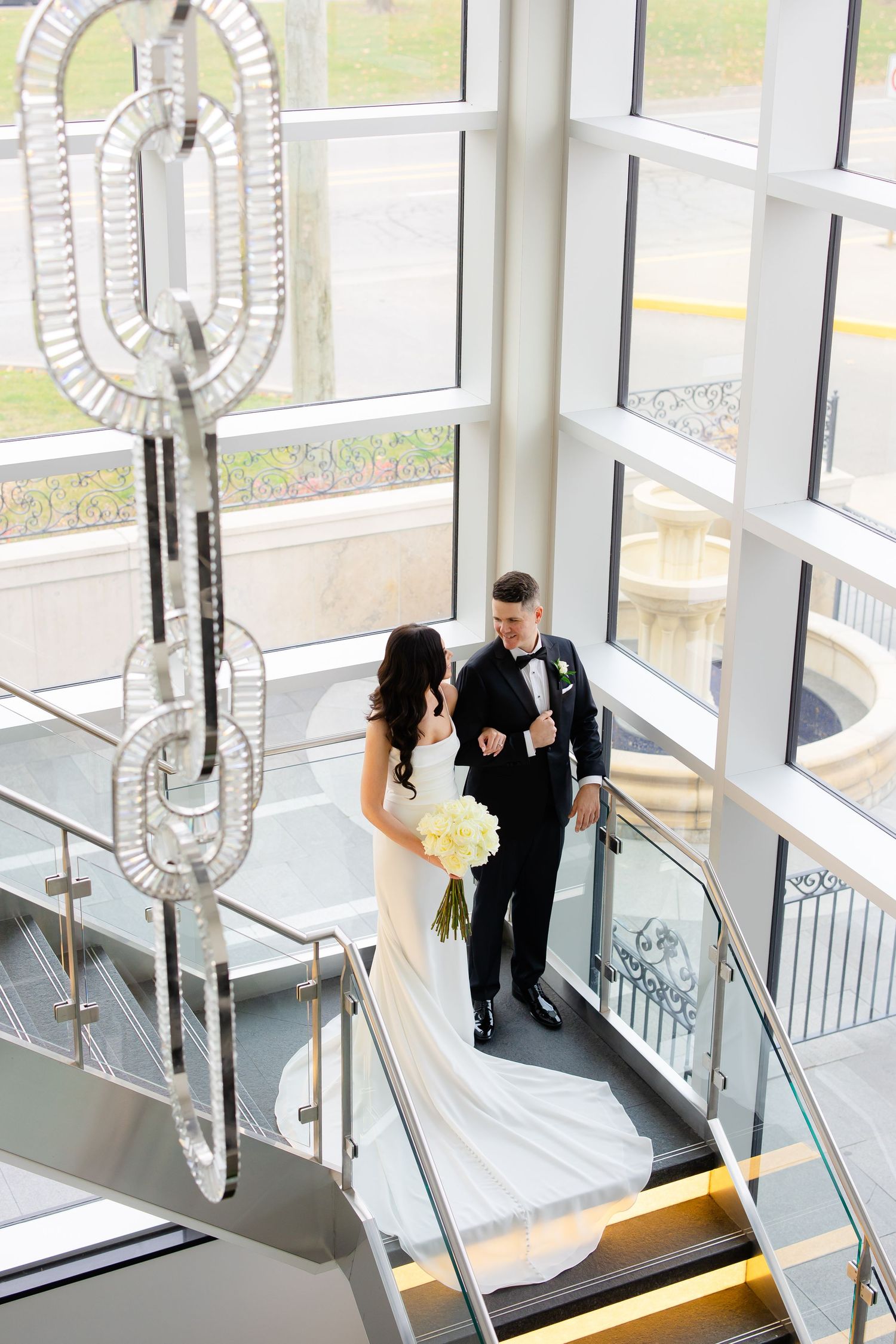 Wedding couple poses on modern staircase with decorative chain chandelier and floor-to-ceiling windows.