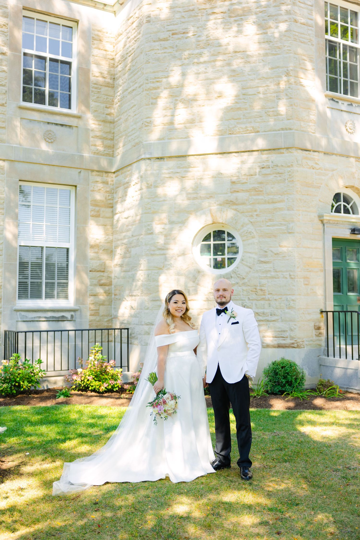 A wedding couple poses against a stone building with a circular window and green door on a sunny day.