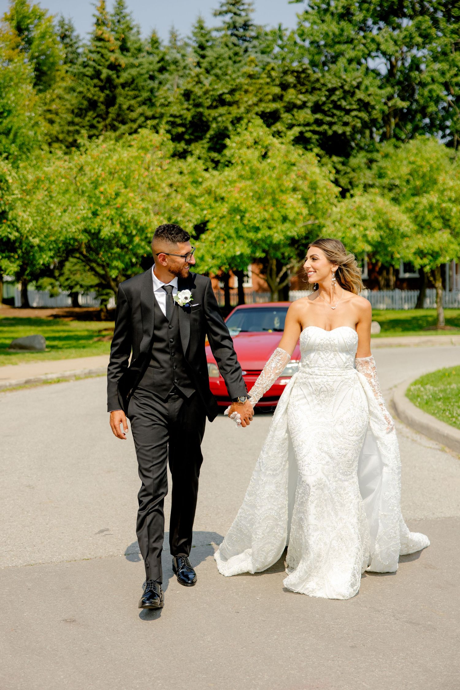 Happy couple strolls hand in hand on their wedding day with a vintage red car in background.