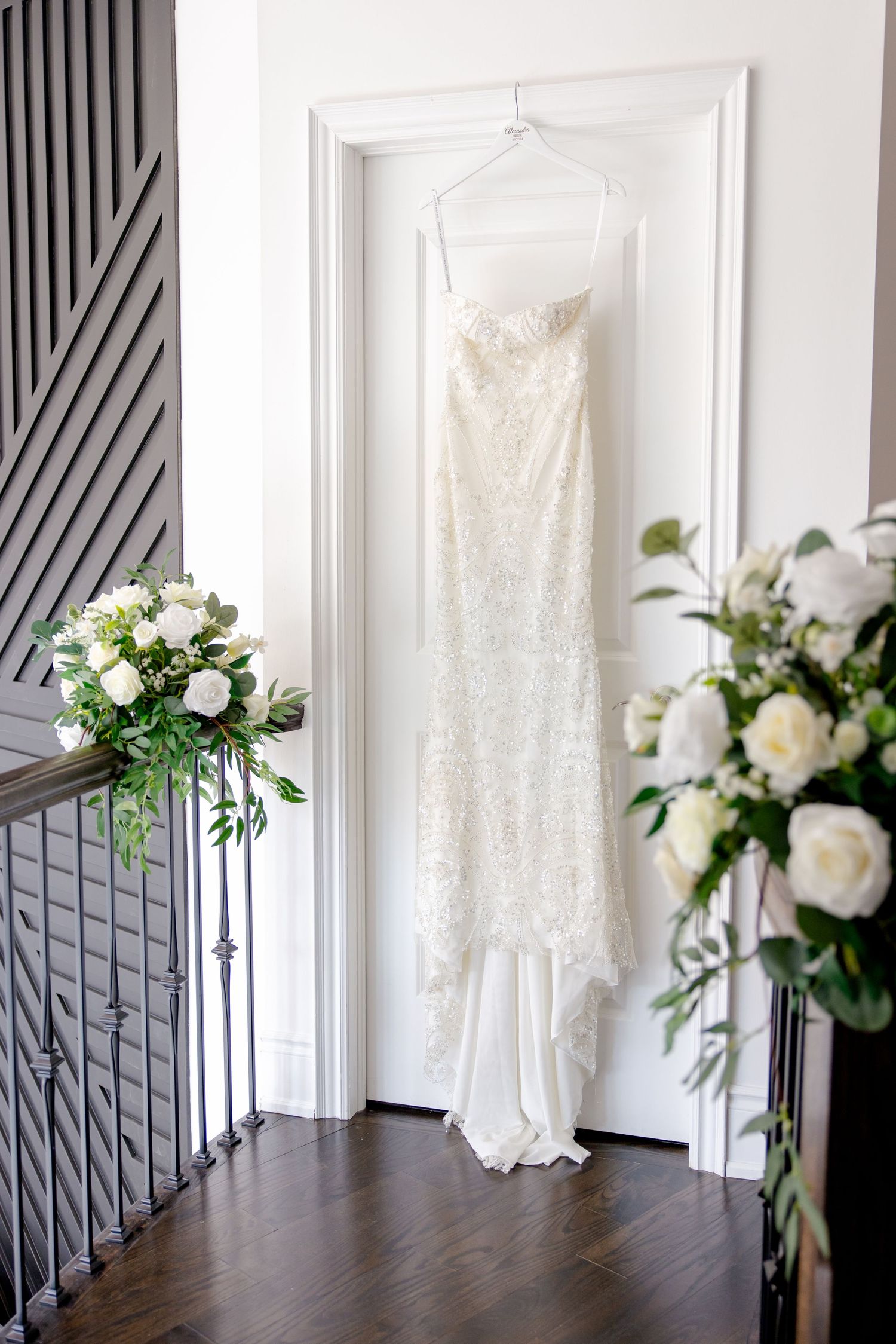 A white lace wedding dress hangs elegantly against a white wall with floral arrangements on either side.