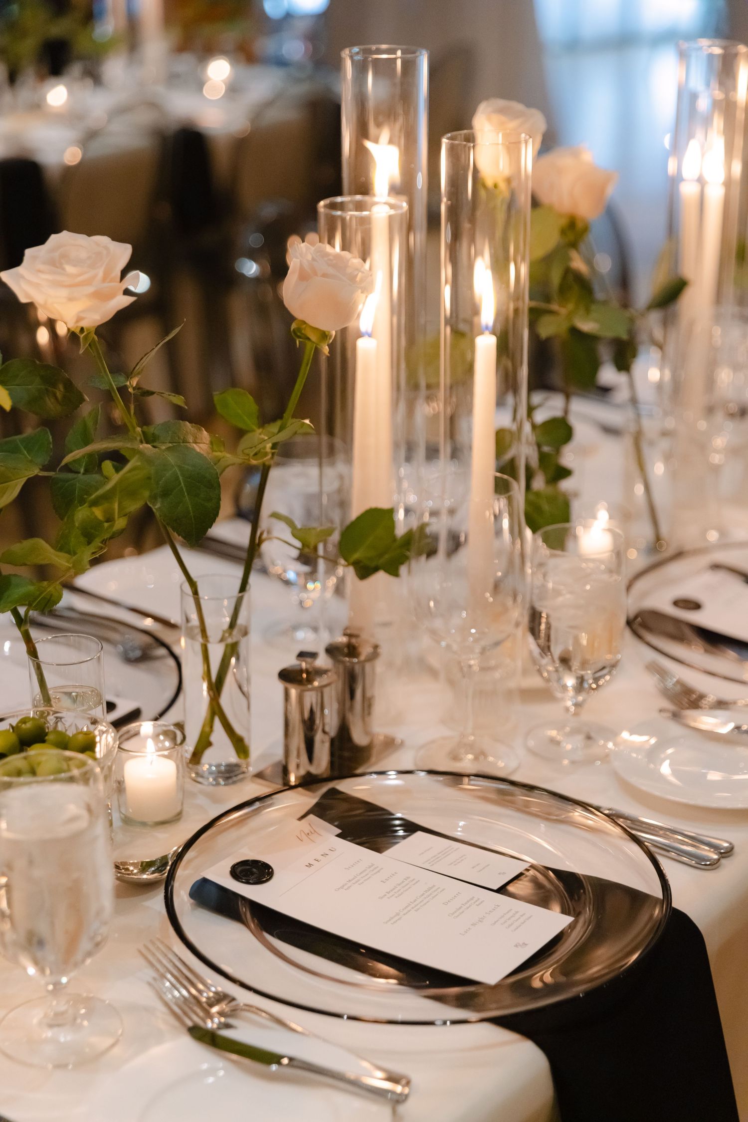 Close-up of formal place setting with glass candleholders, white roses, and black napkin accents.