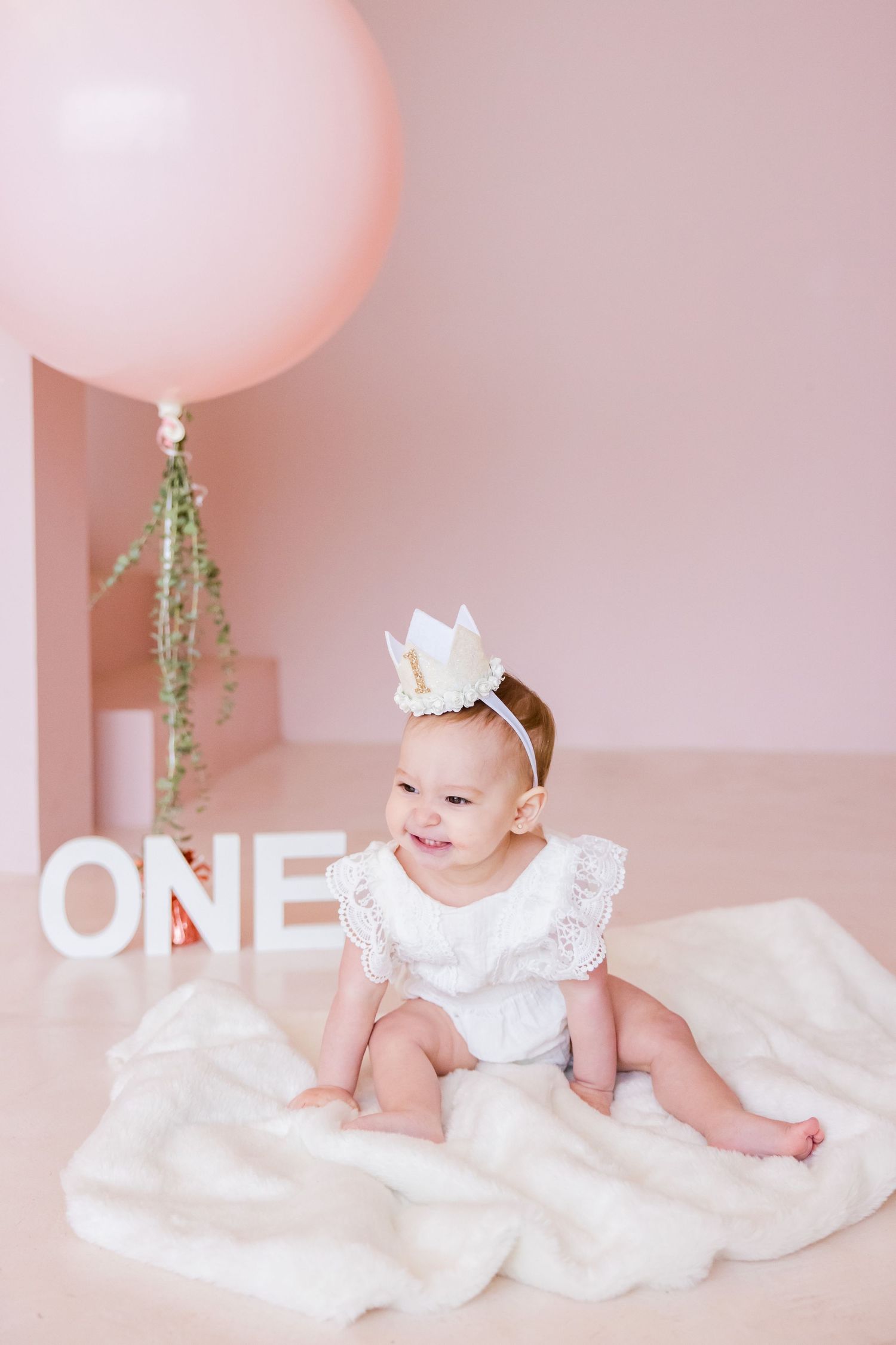 Birthday photo with white dress, crown and pink balloon against soft pink background.
