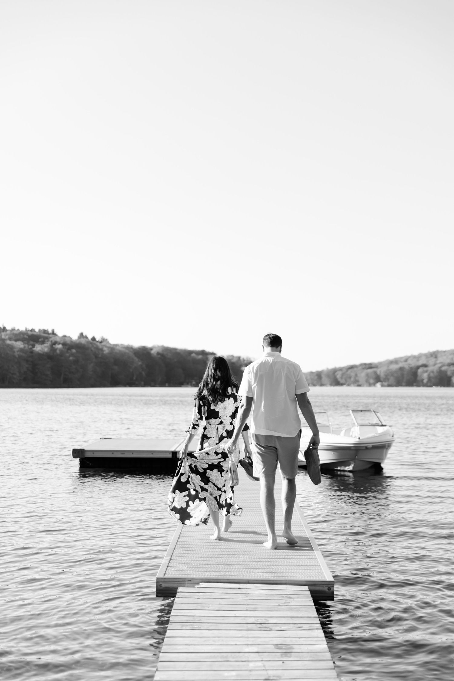 Black and white photo of couple walking on wooden dock with boat in background.