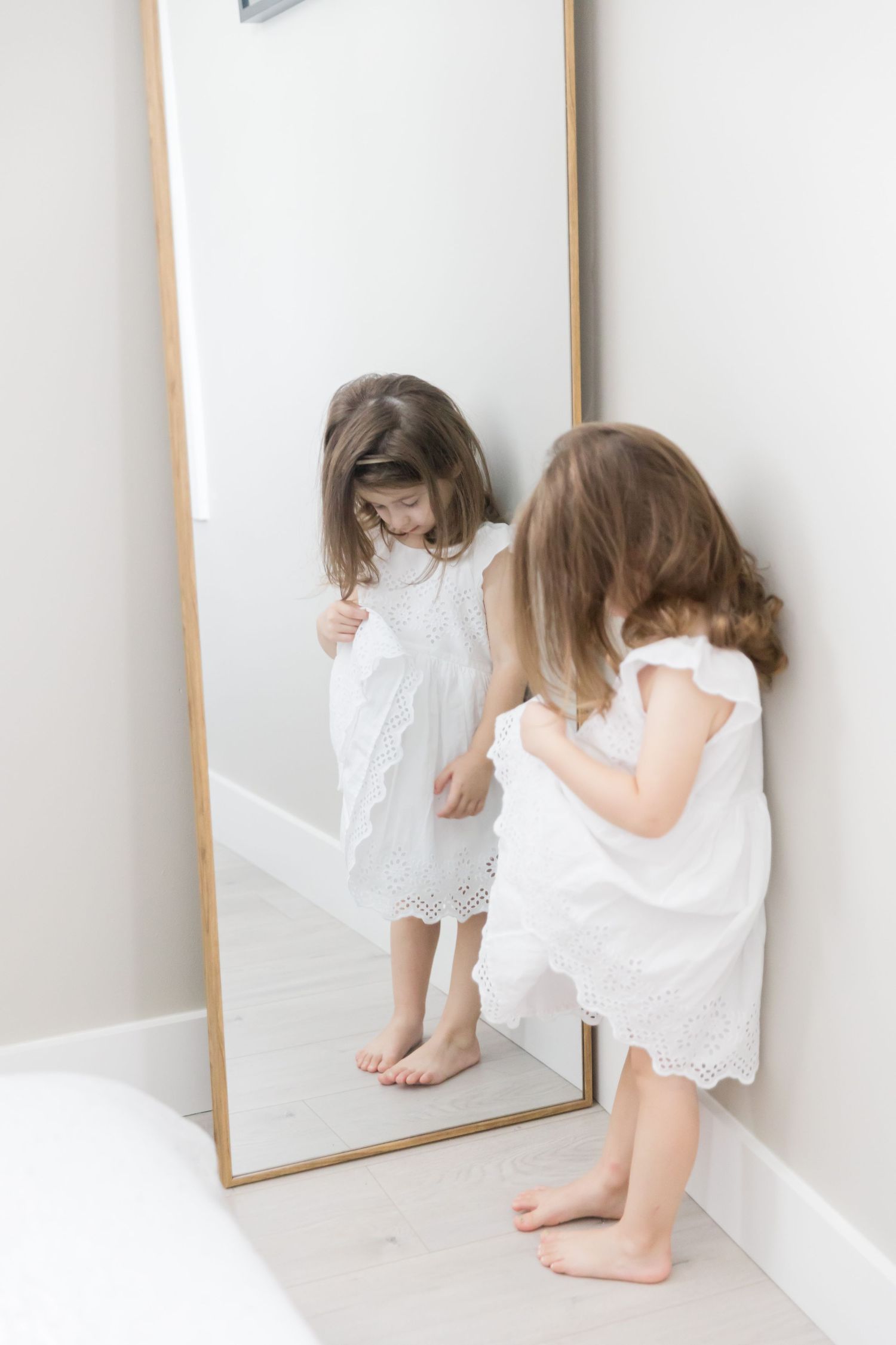 Young child in white dress looks at reflection in full-length mirror against white wall.