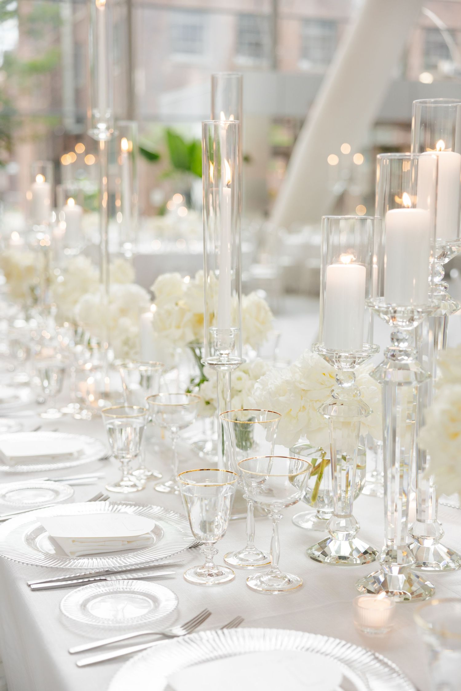 Modern white wedding reception table with tall glass candleholders and fresh white floral arrangements.