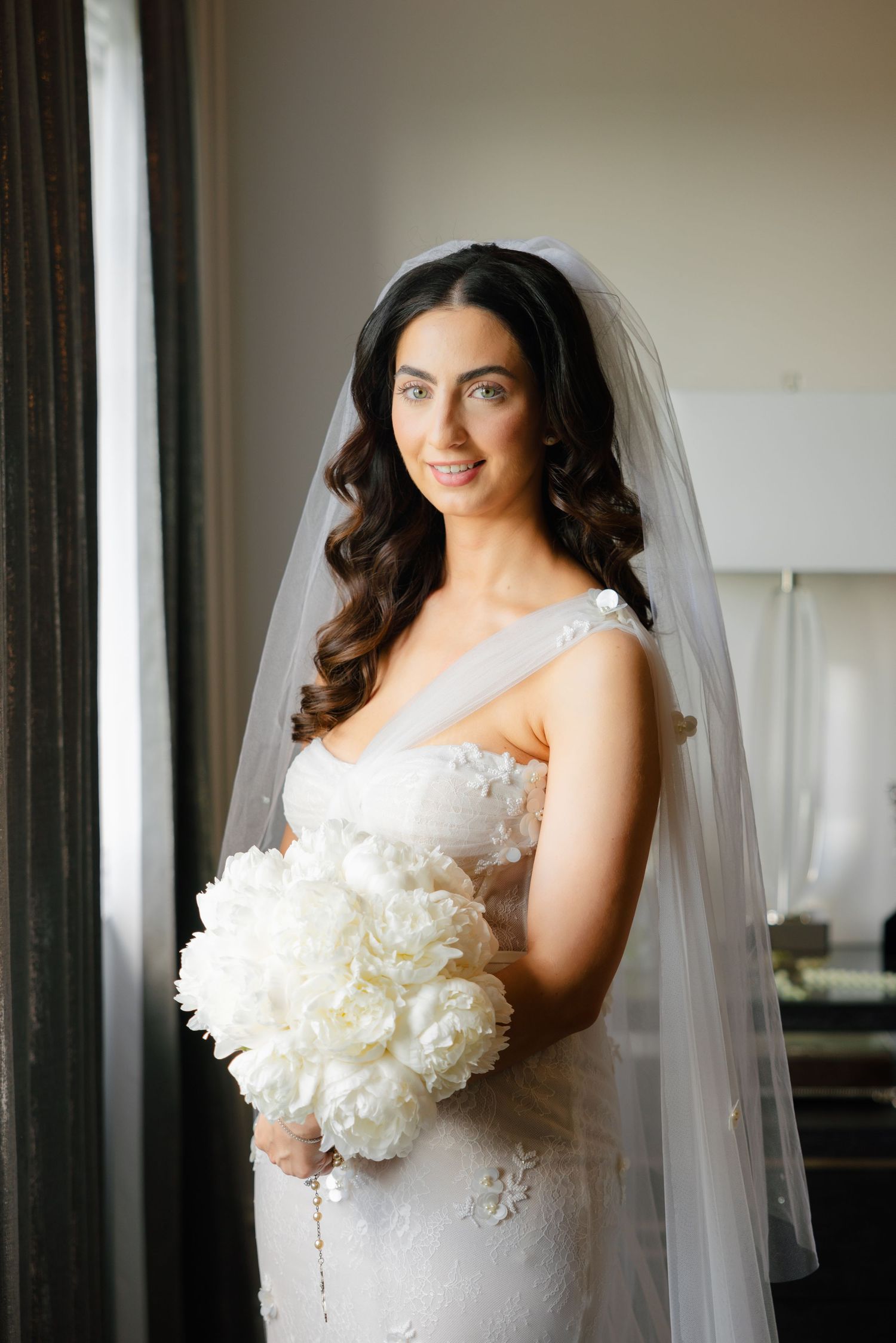 Natural window light illuminates bride with flowing veil and classic white peony bouquet.