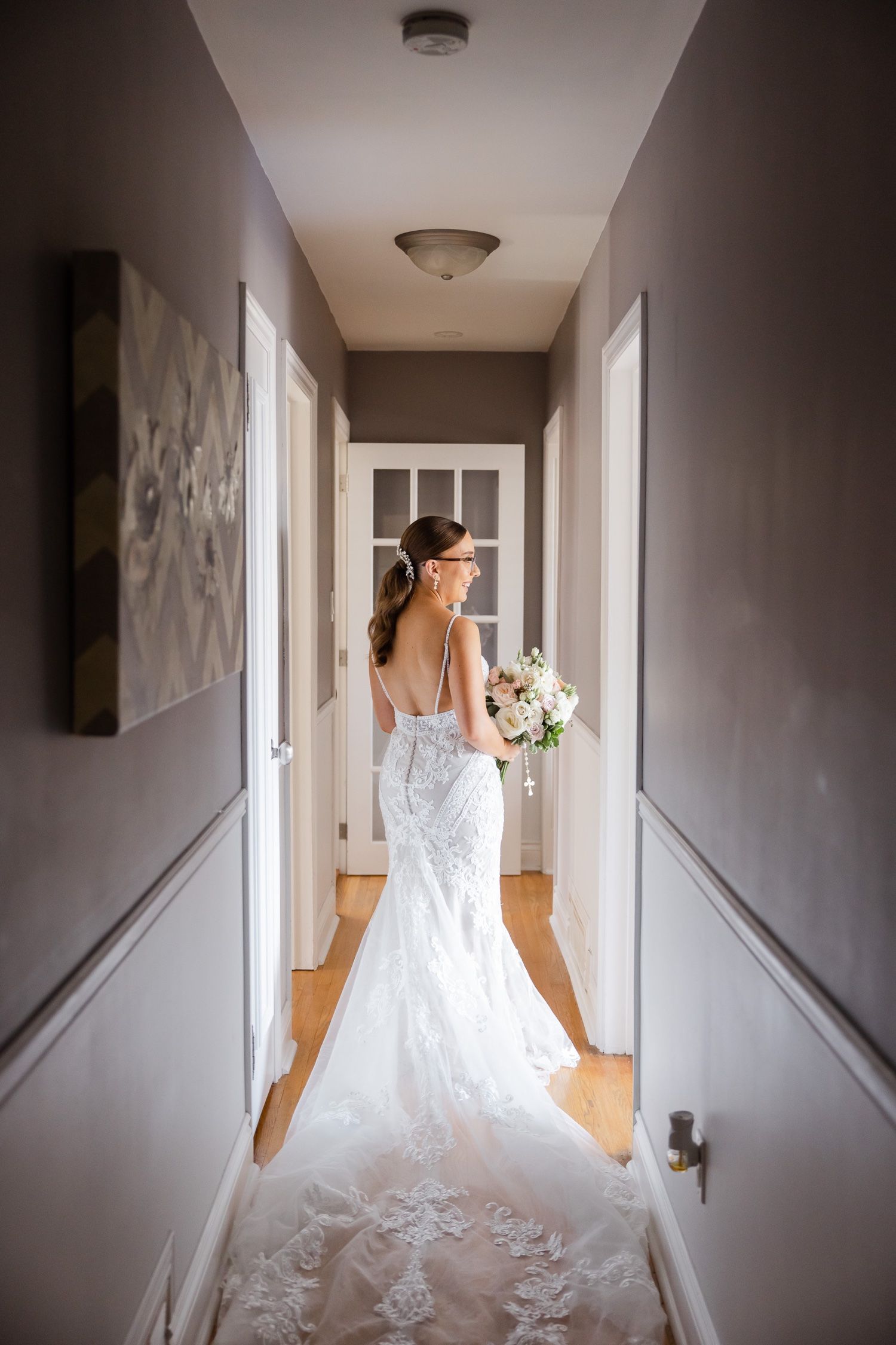 A bride in a lace wedding gown walks down a hallway holding a bouquet.