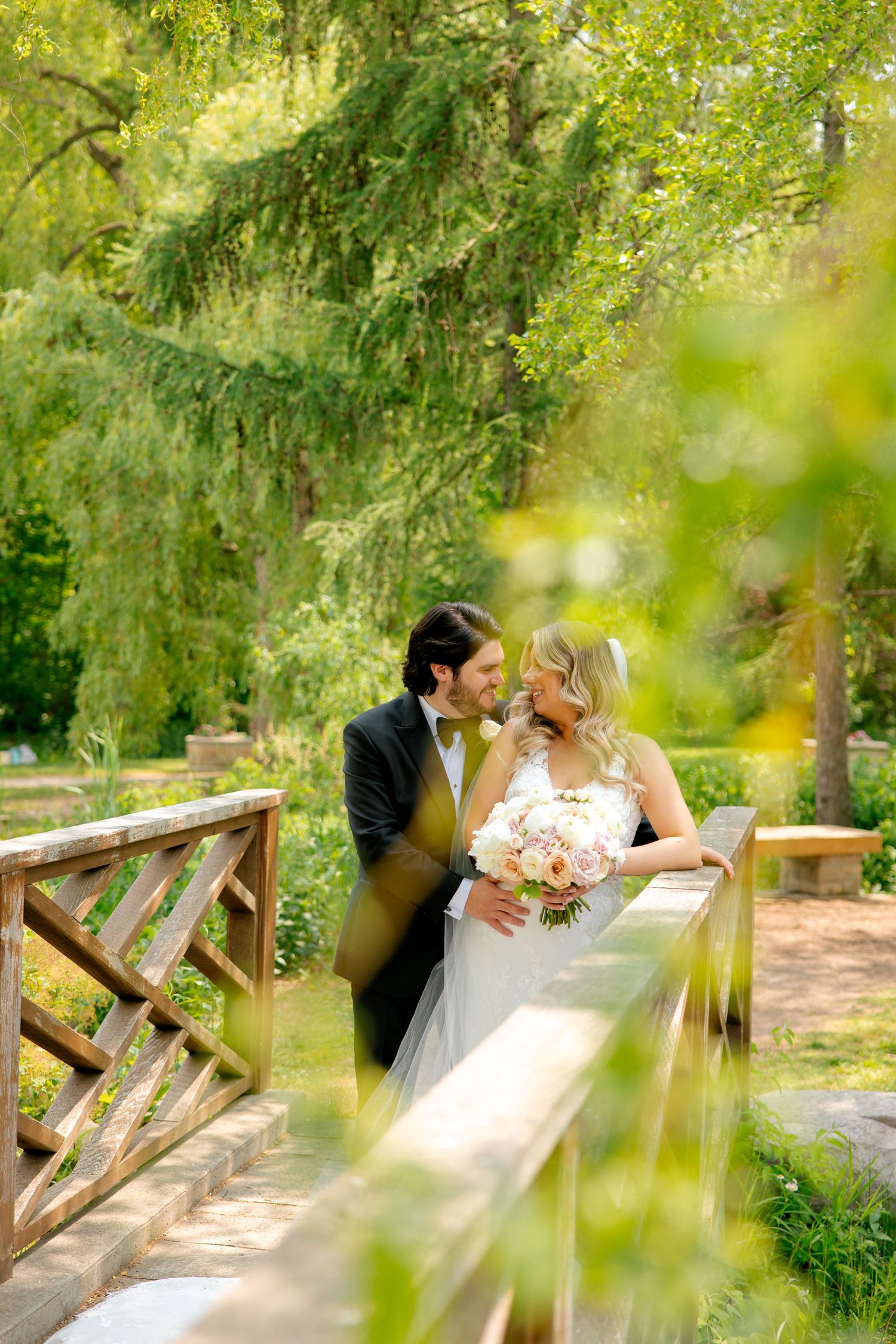 Wedding couple shares intimate moment on wooden bridge in garden setting with soft natural lighting.