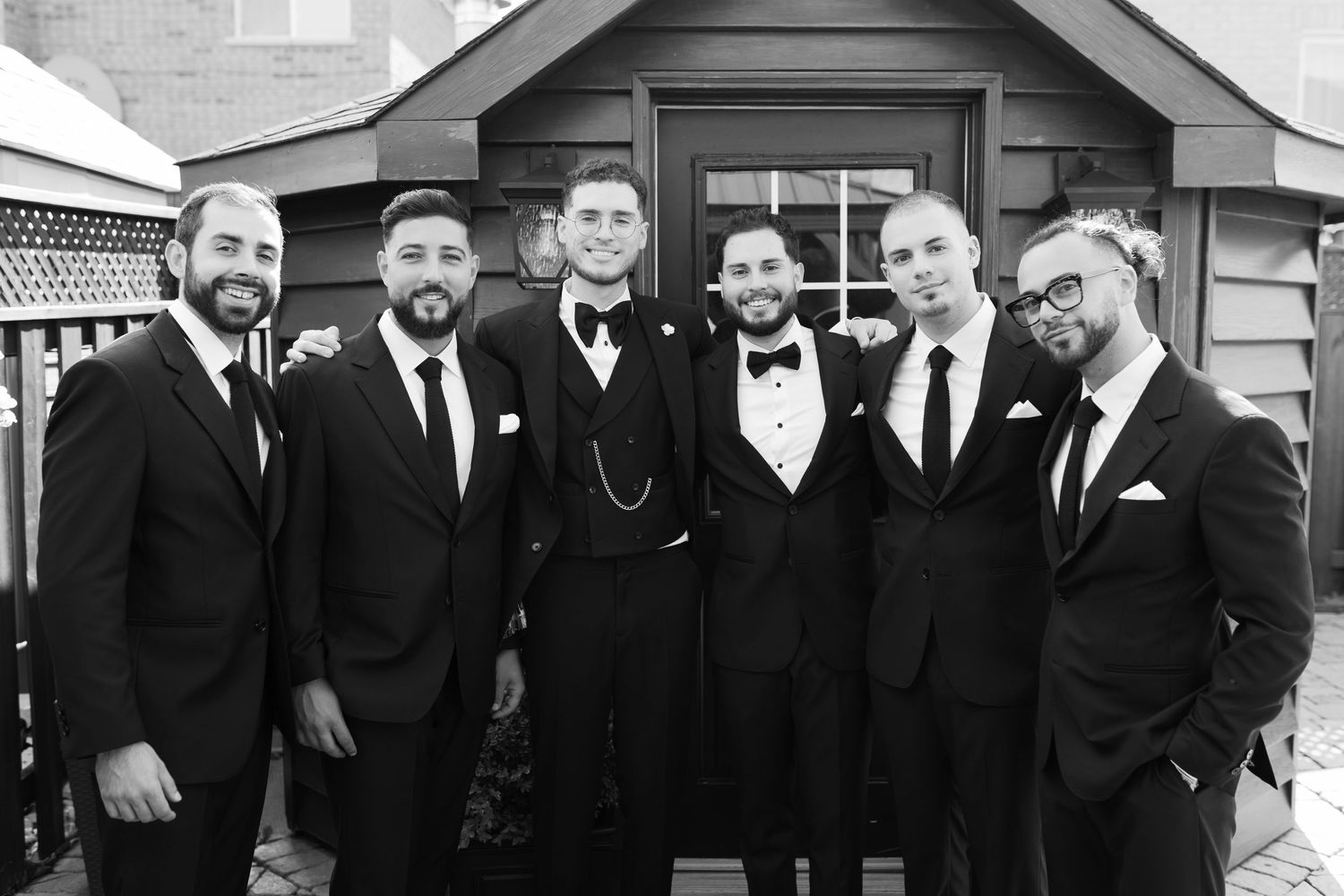 A formal black and white group photo of groomsmen outside a wooden structure.