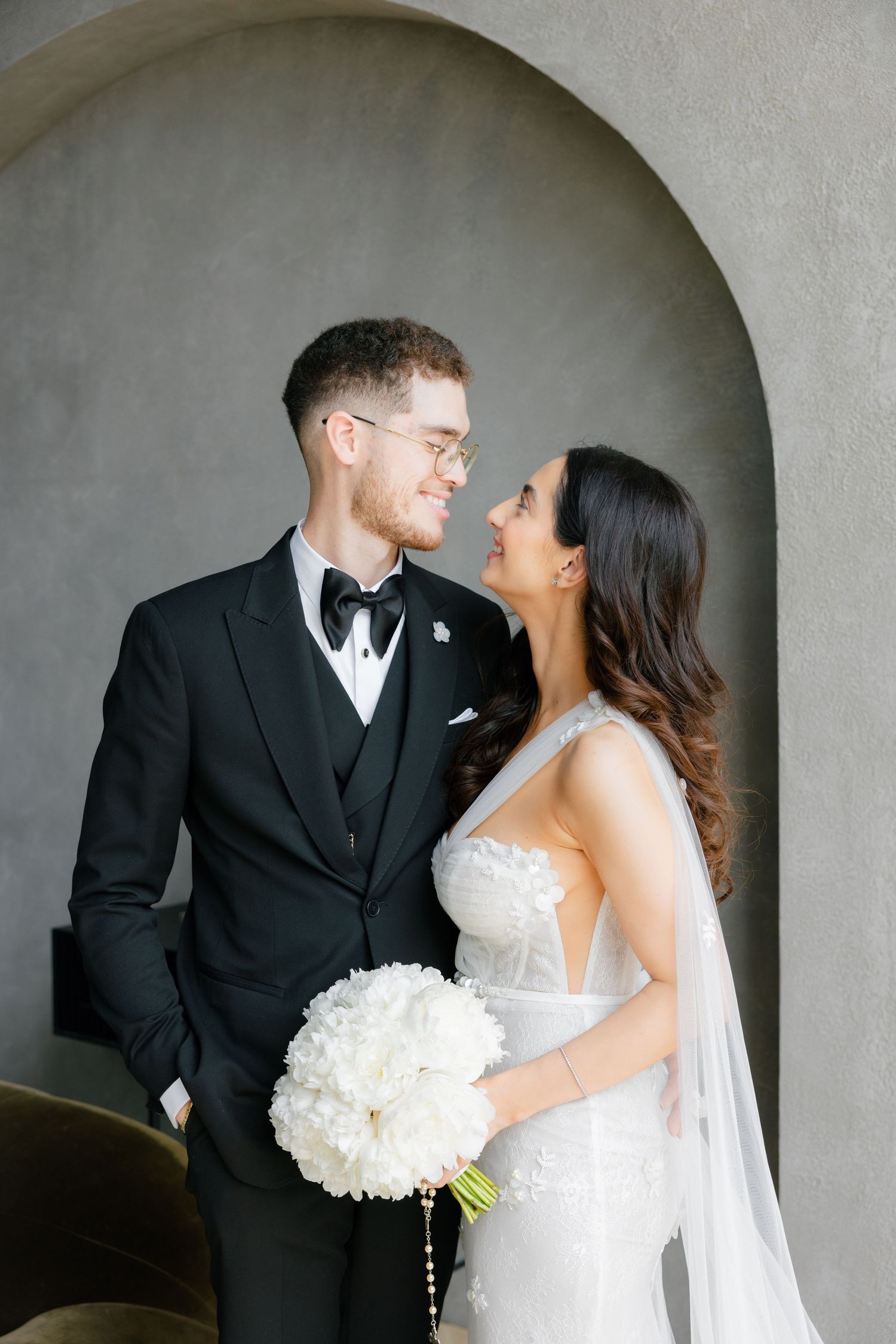Newlyweds share joyful moment together while posing with white bouquet under arch.