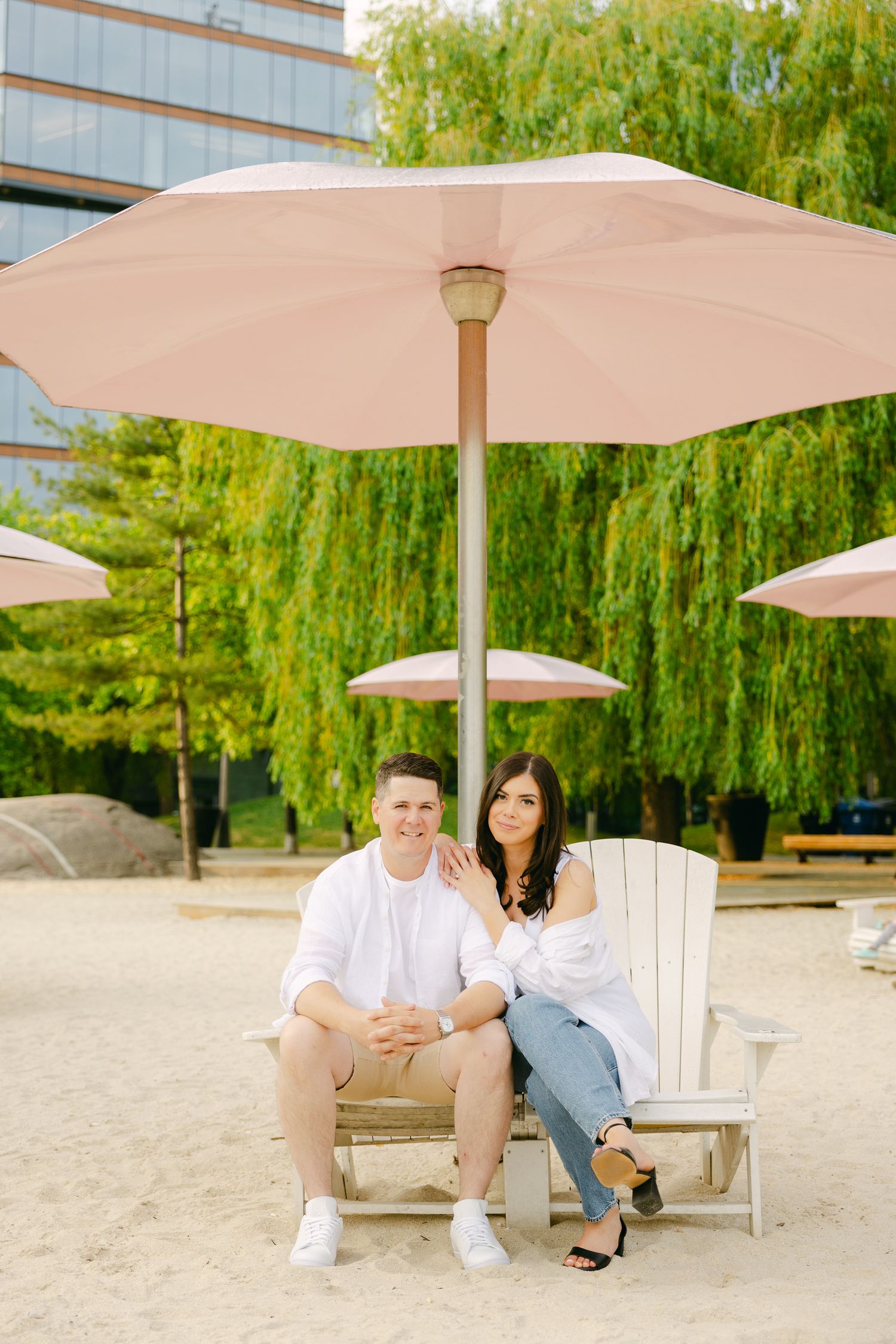 Couple sitting under pink umbrella with willow trees in background.