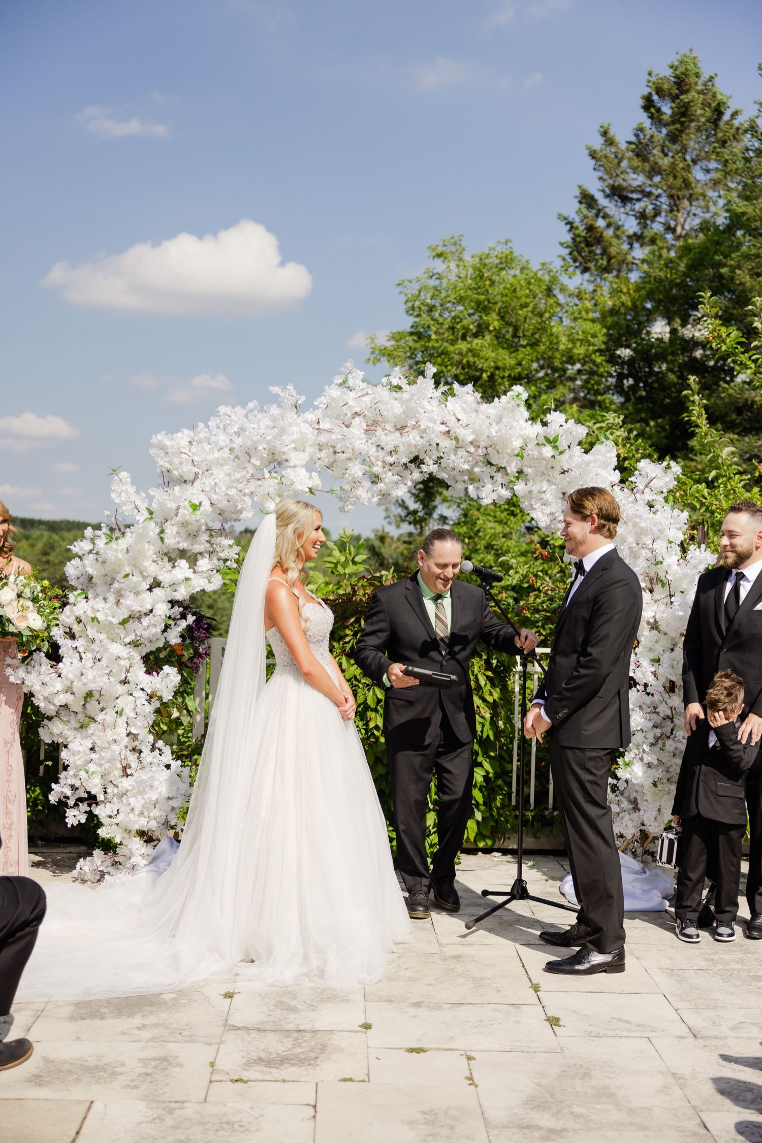 Wedding ceremony taking place beneath an elaborate white floral arch on a stone patio.