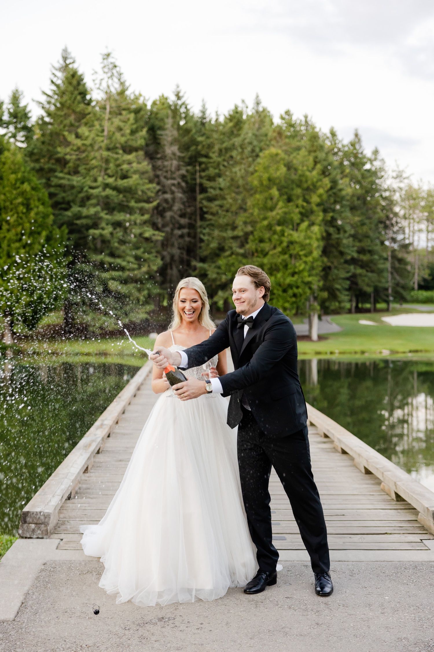 A couple pops champagne during their wedding photoshoot on a lakeside dock with evergreen trees in the background.