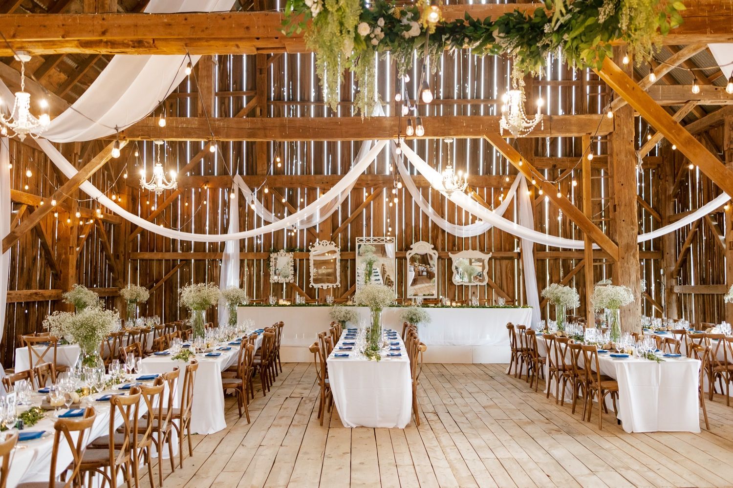 A rustic barn wedding reception setup with white draping, wooden tables, and elegant table settings.