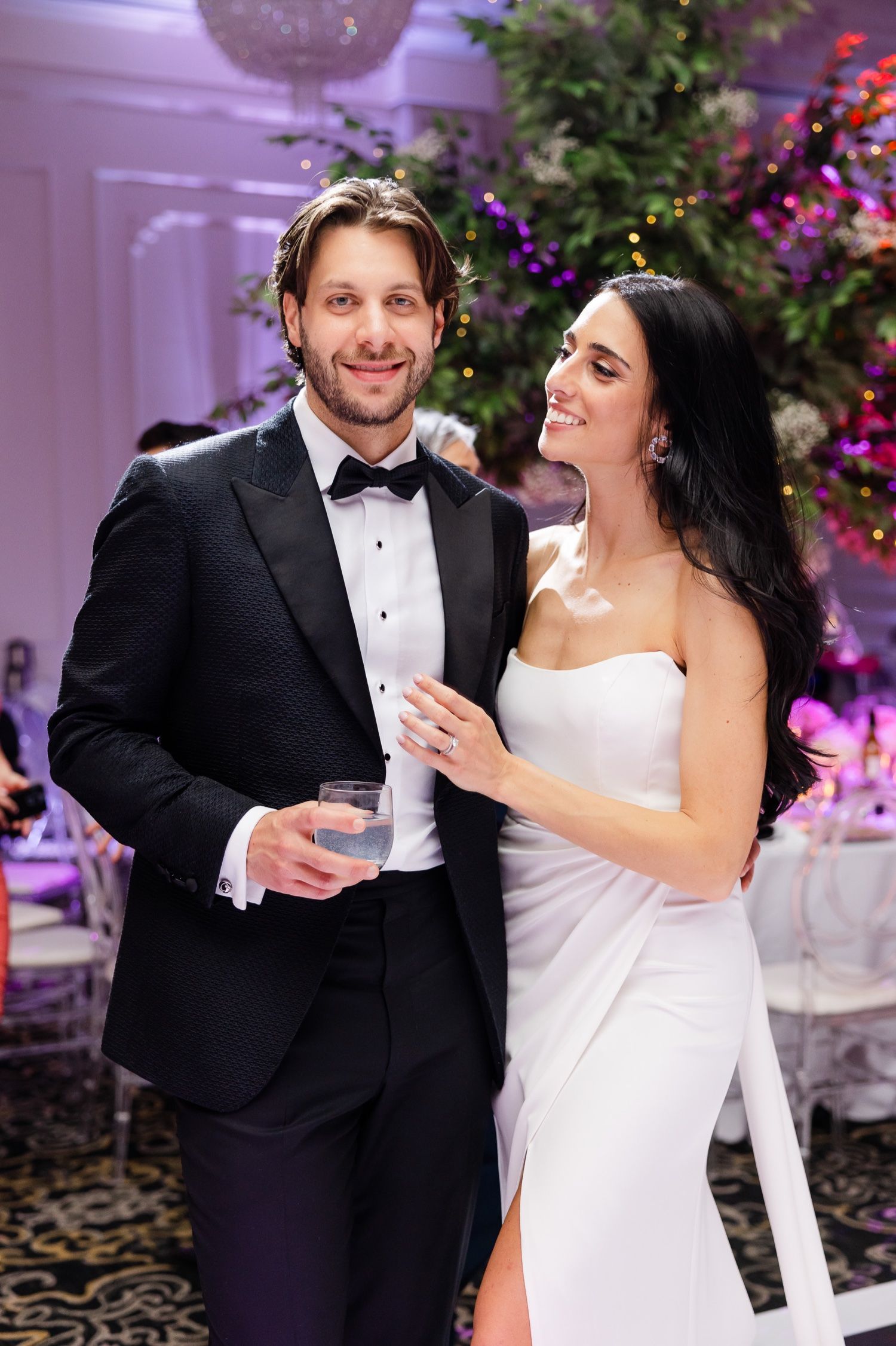 Couple shares drinks and laughter at their wedding reception under purple mood lighting.