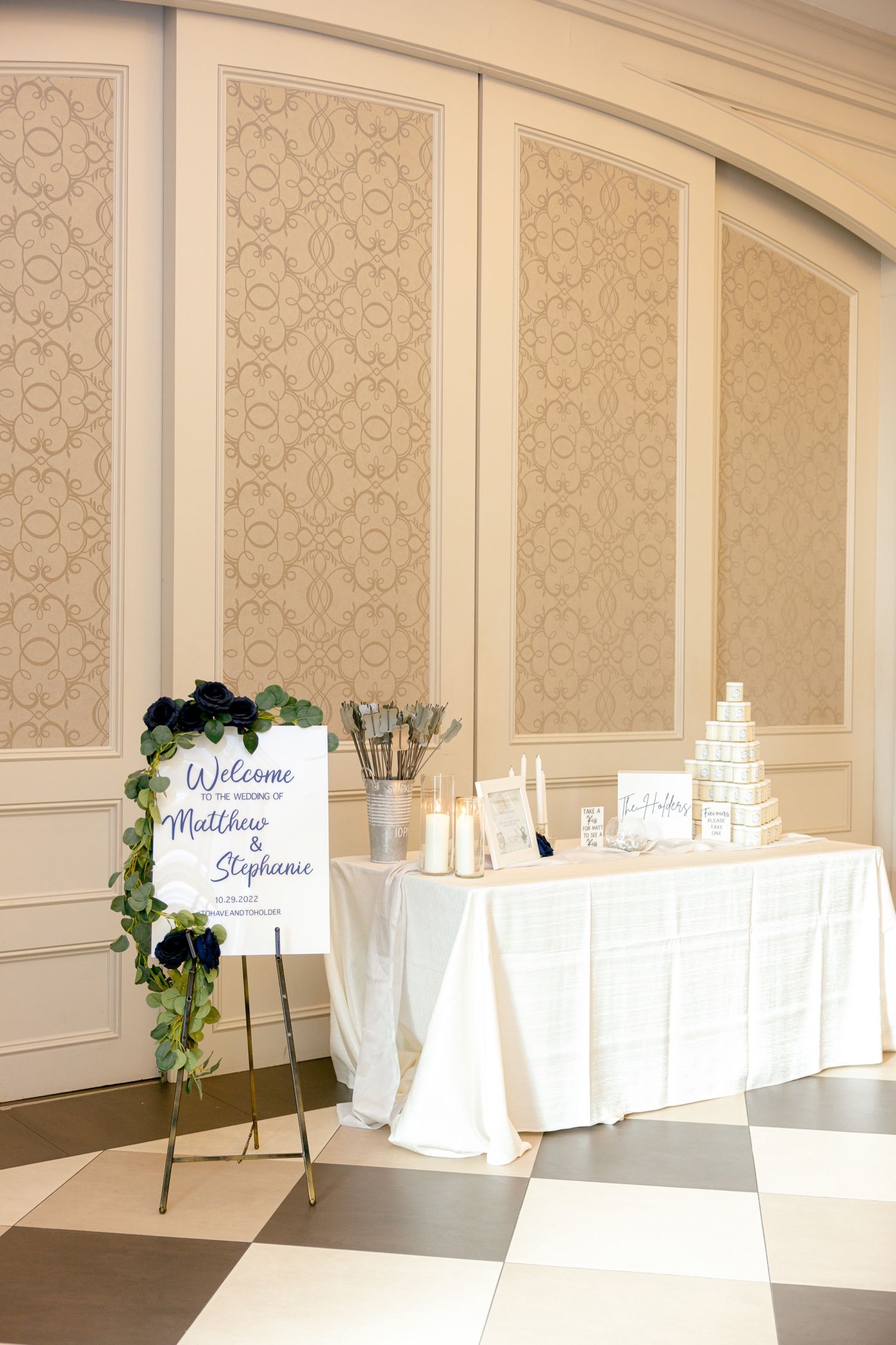 Welcome sign and display table at wedding reception with cream wallpaper panels and checkered floor.