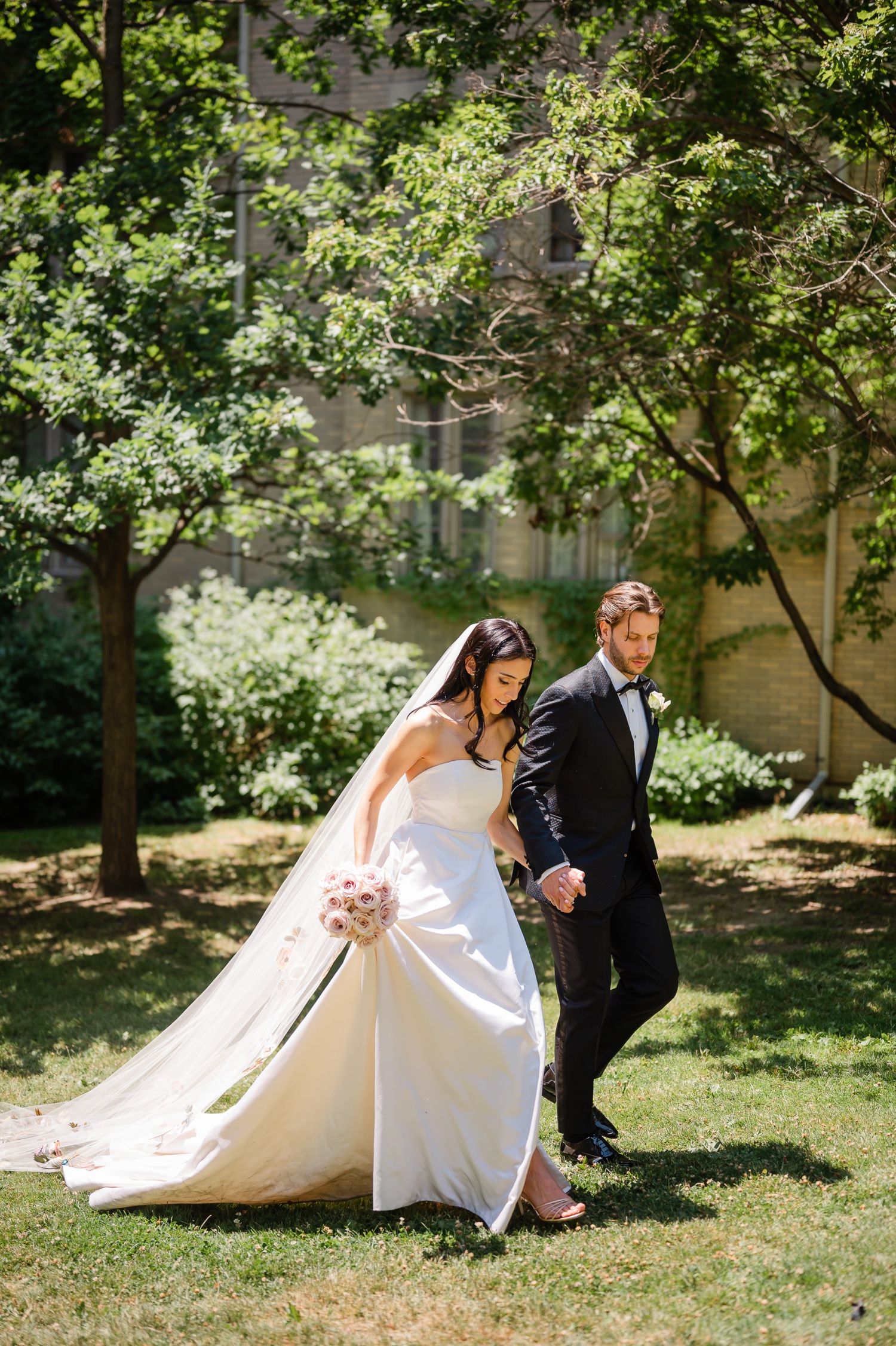 Wedding couple walking through a sunlit garden with the bride's detailed train flowing behind.