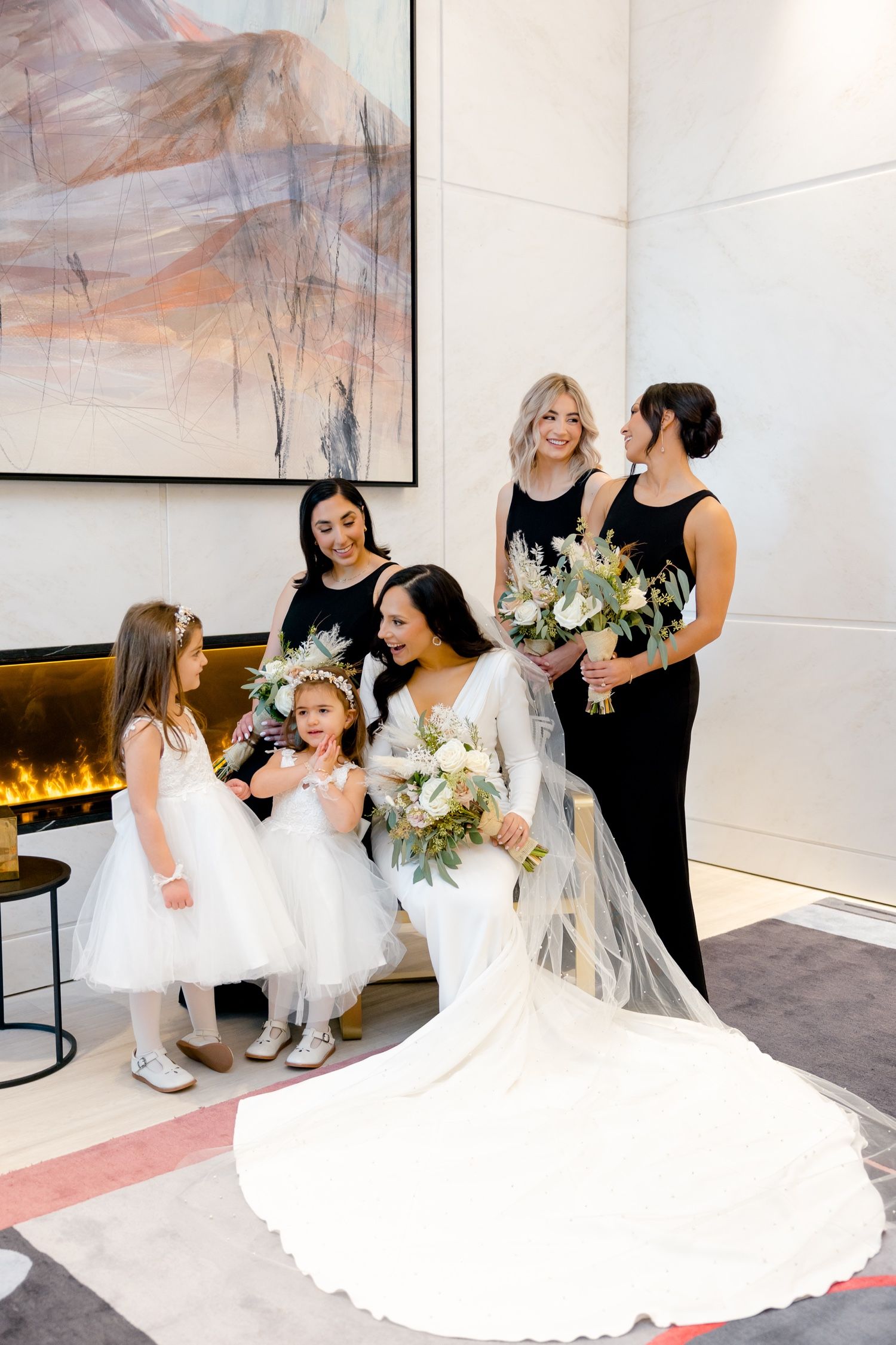 Bridal party gathers around bride in flowing white gown beneath modern abstract artwork.