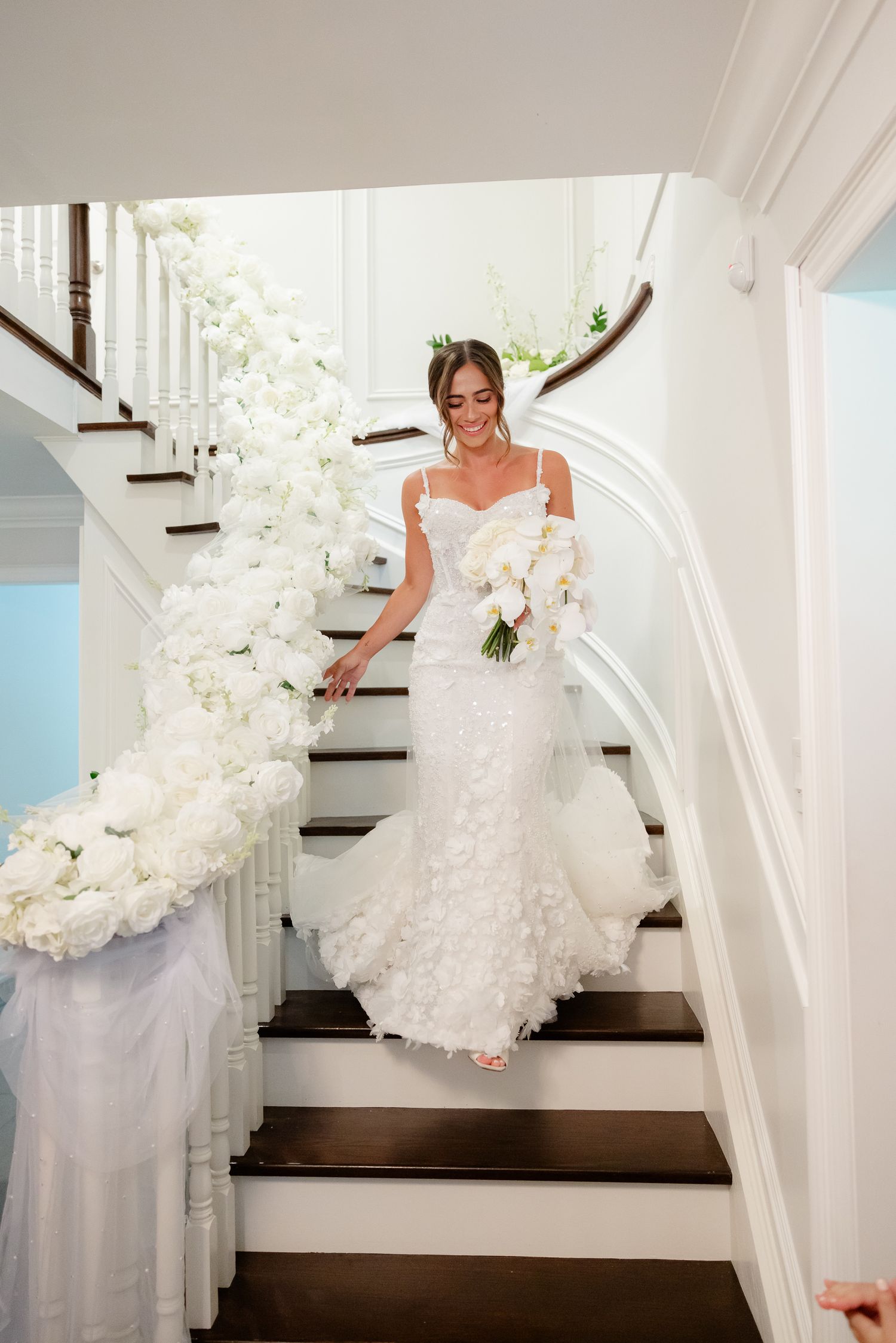 Bride descends elegant white staircase decorated with cascading white orchids, wearing fitted lace gown with bouquet.