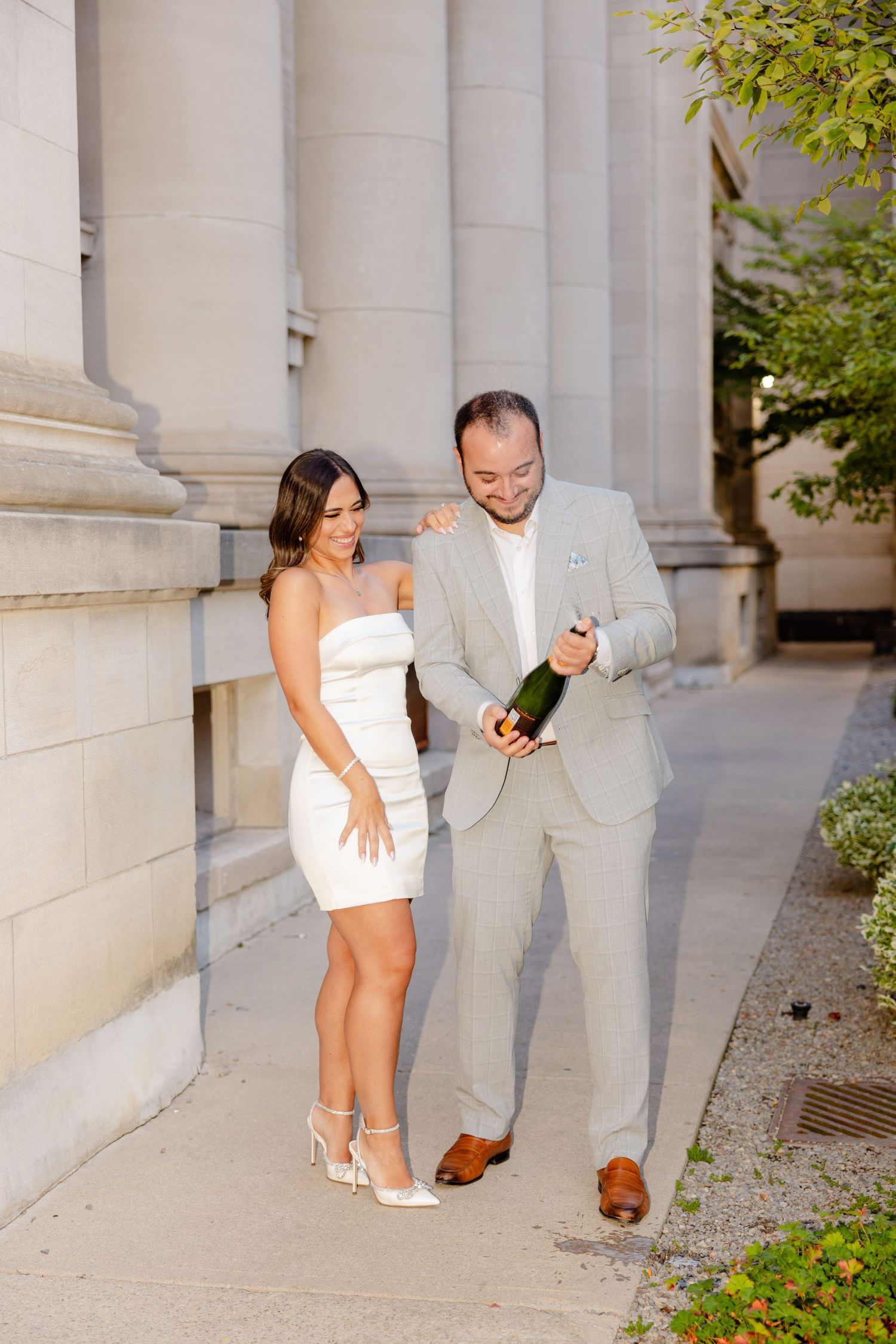 Stylish couple in white dress and light gray suit poses with champagne by stone columns.