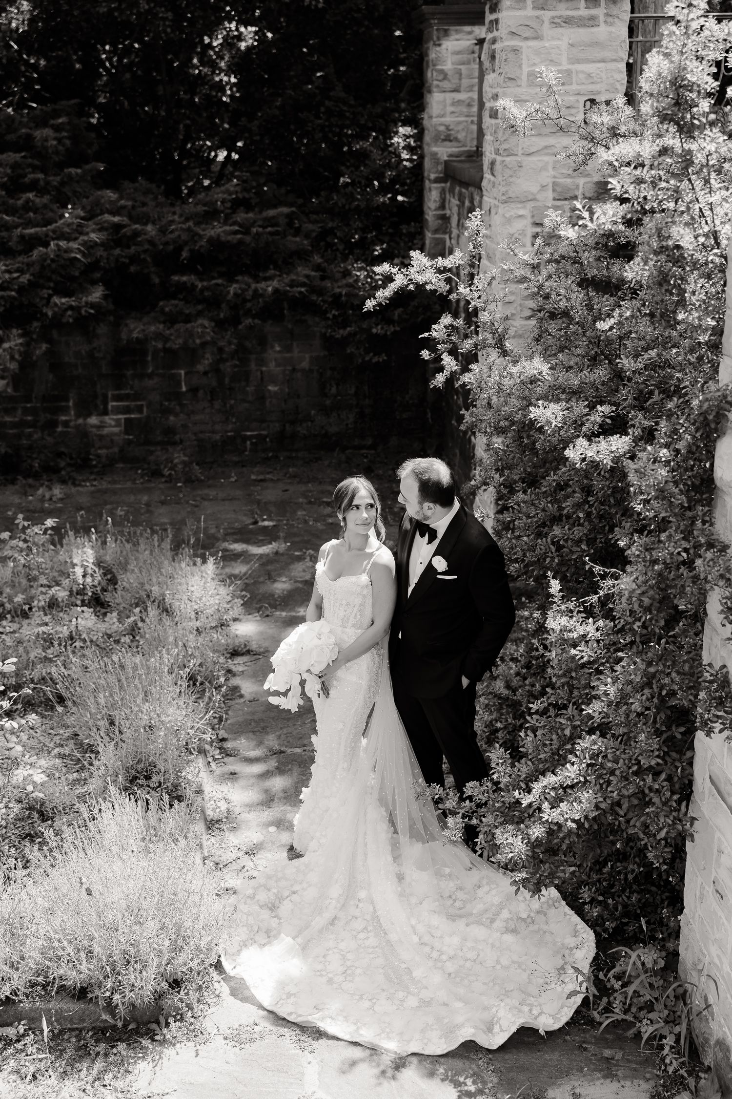 A bride and groom pose together in a black and white photograph against a stone wall and garden setting.