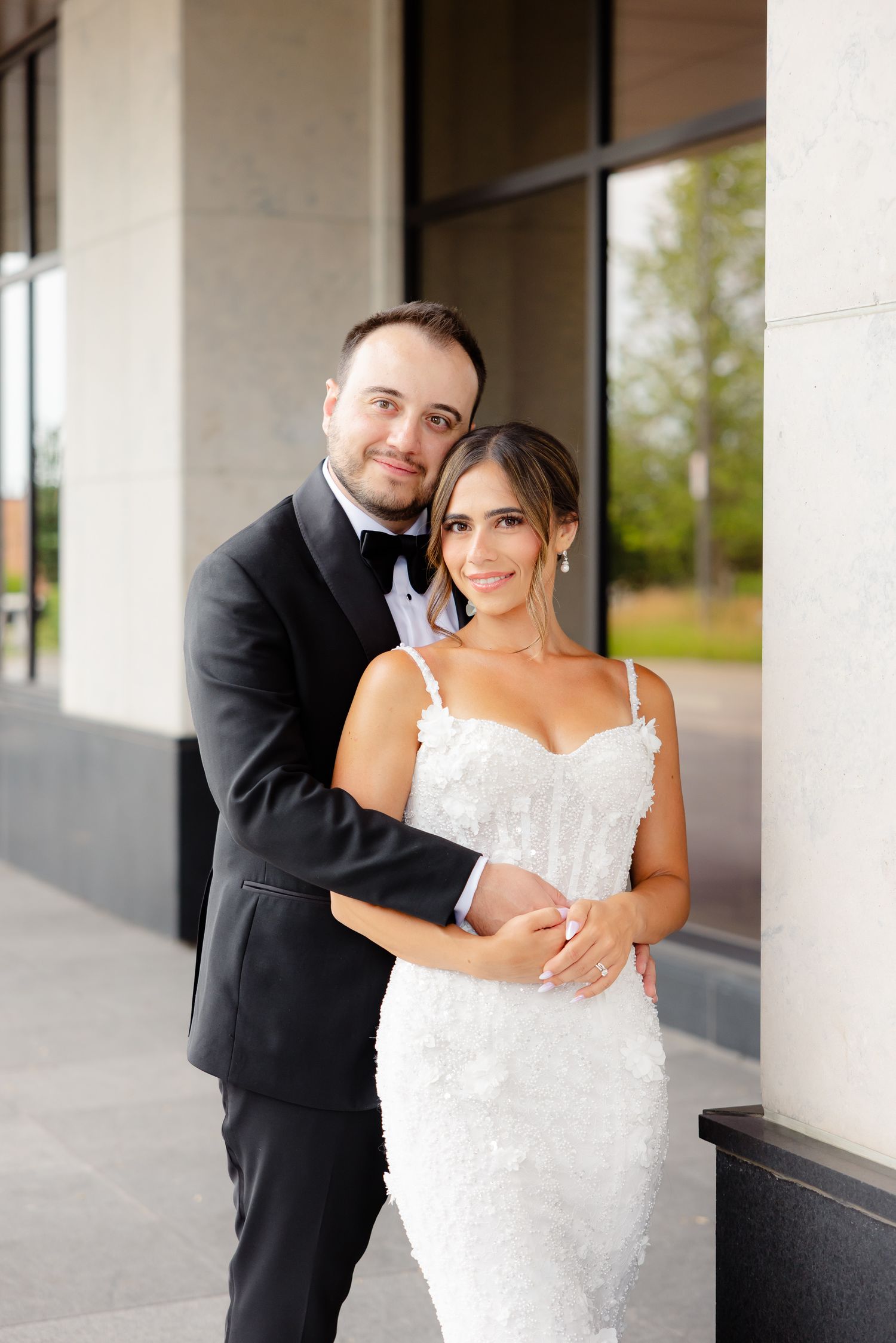 A couple embraces in formal wedding attire during their wedding photoshoot outside a modern building.