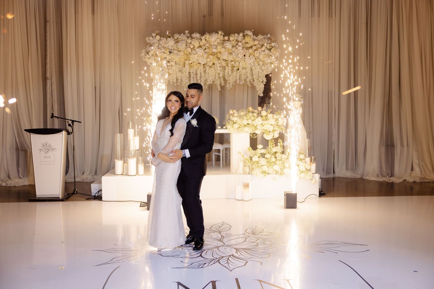 Elegant wedding dance pose with sparkler effects and white floral backdrop on monogrammed floor.