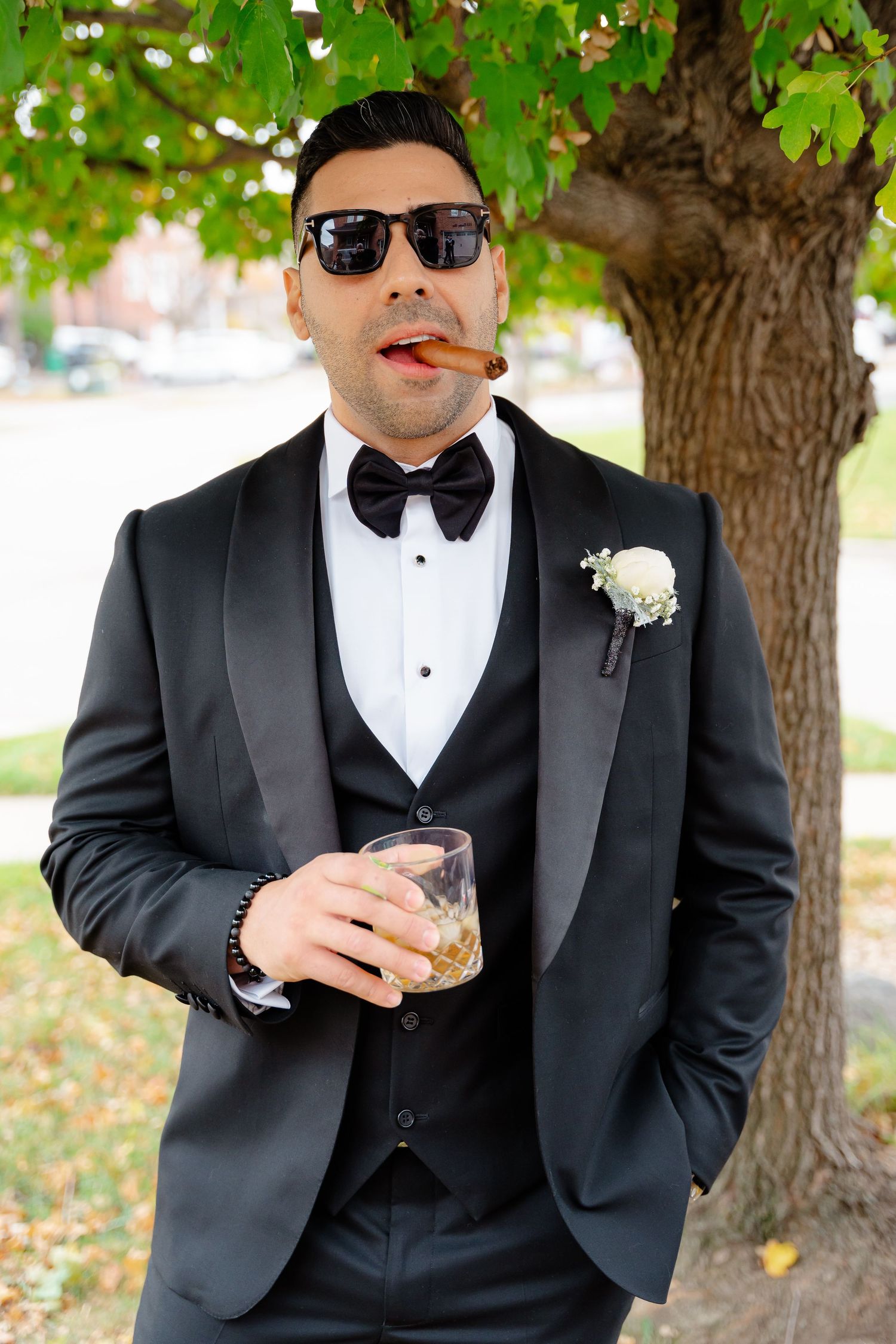 Sharp dressed wedding party member in tuxedo enjoying a cigar and drink under a tree.