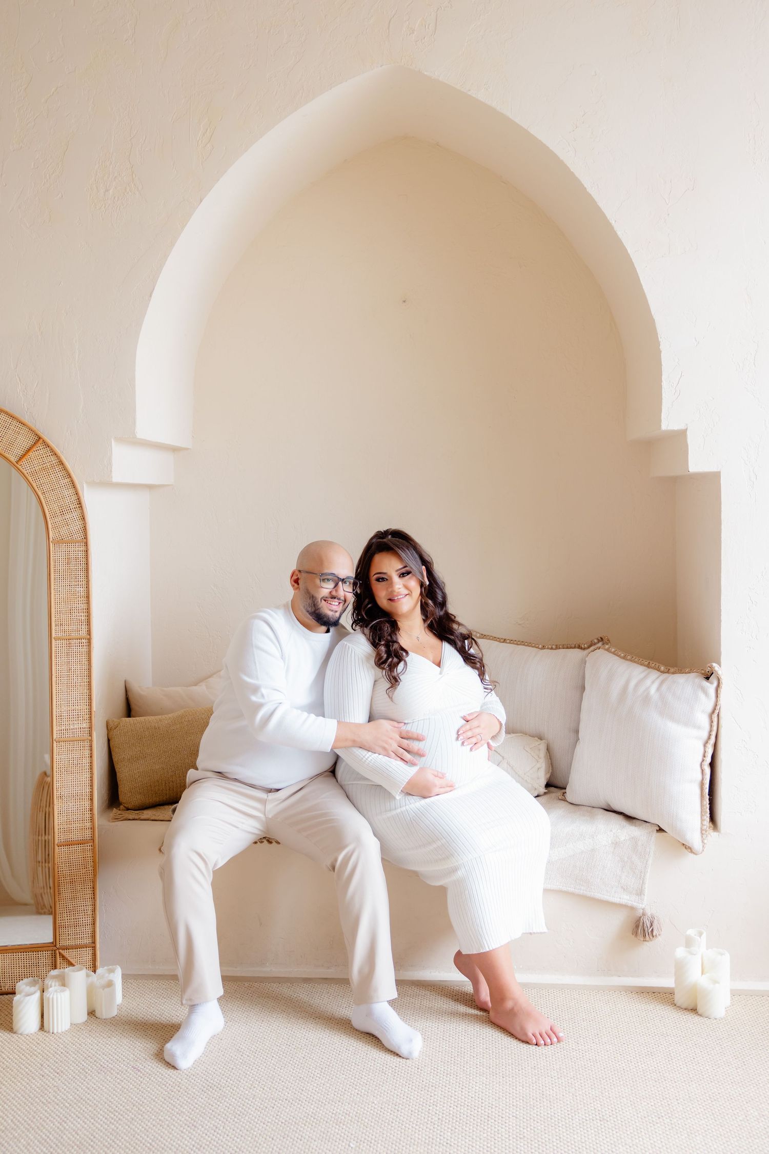 A romantic couple in all white sit together in an arched alcove decorated with candles and an ornate mirror.