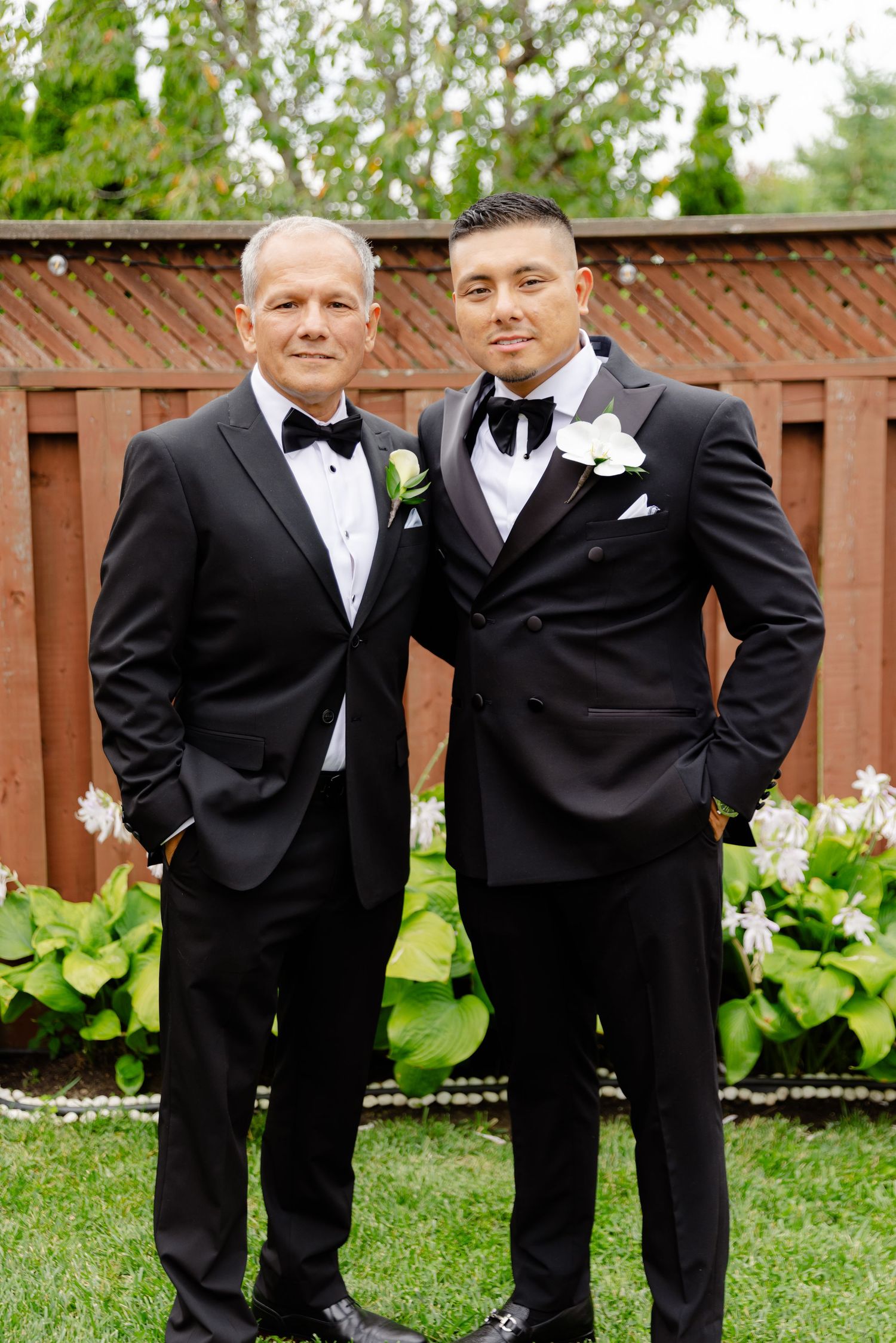 Wedding party members in black tuxedos pose together in front of a wooden fence and garden setting.