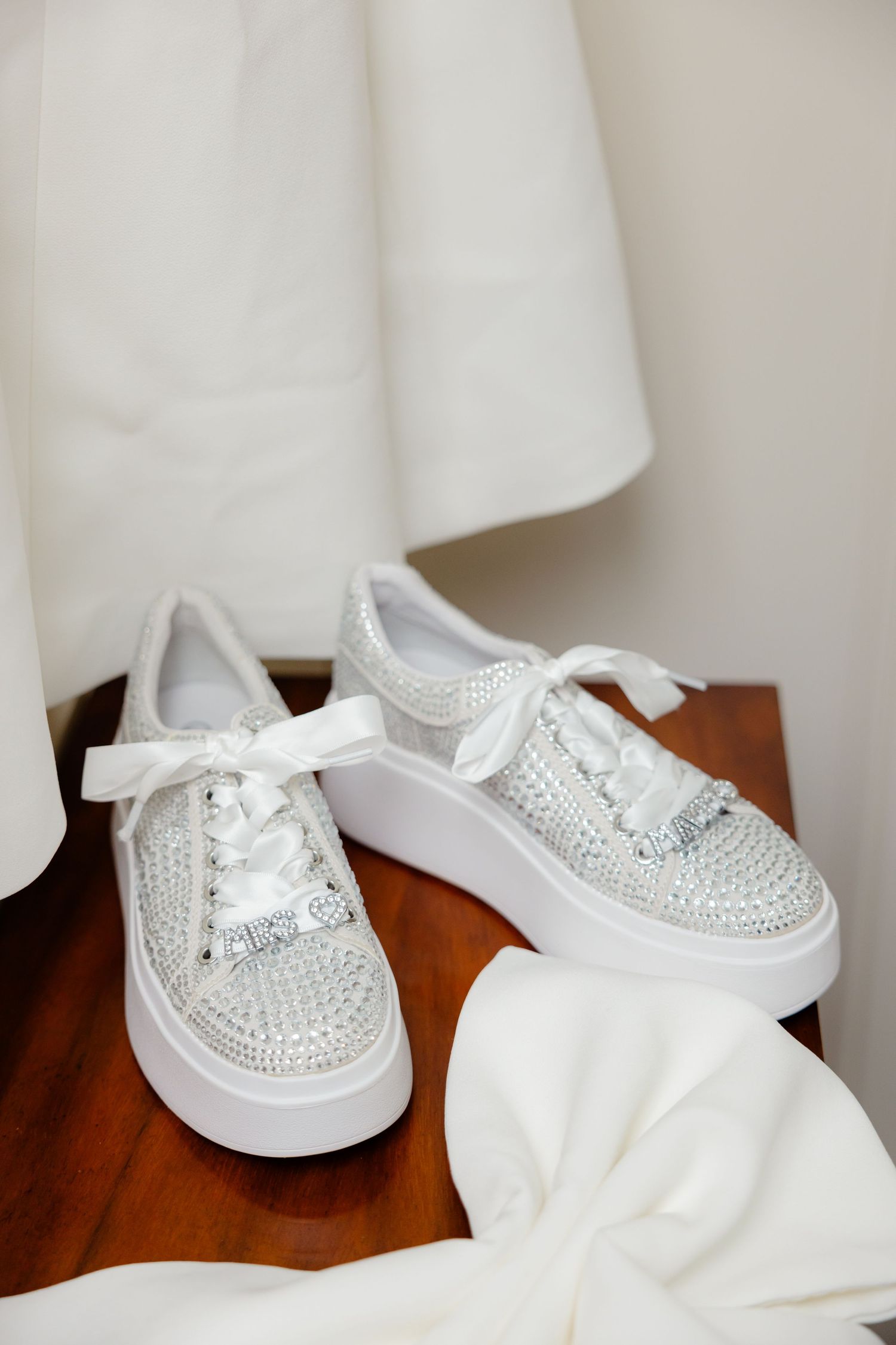 Crystal-embellished white sneakers displayed on dark wood table with white fabric.
