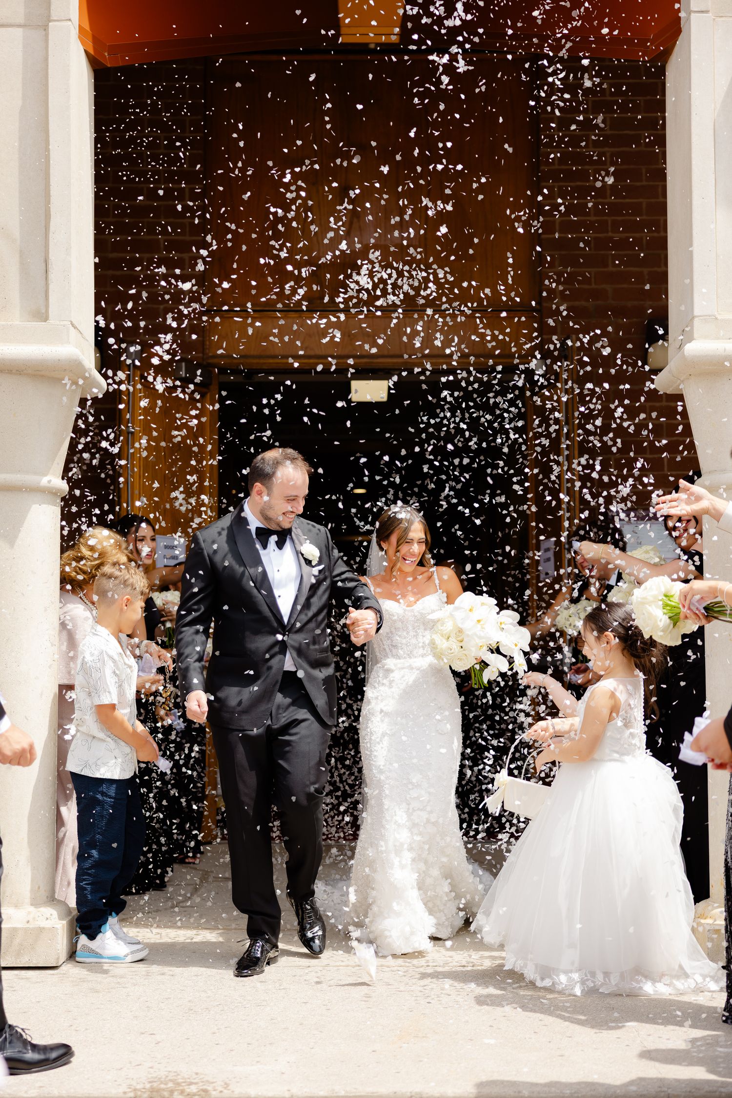 Newlywed couple exits church while guests shower them with white confetti on their wedding day.