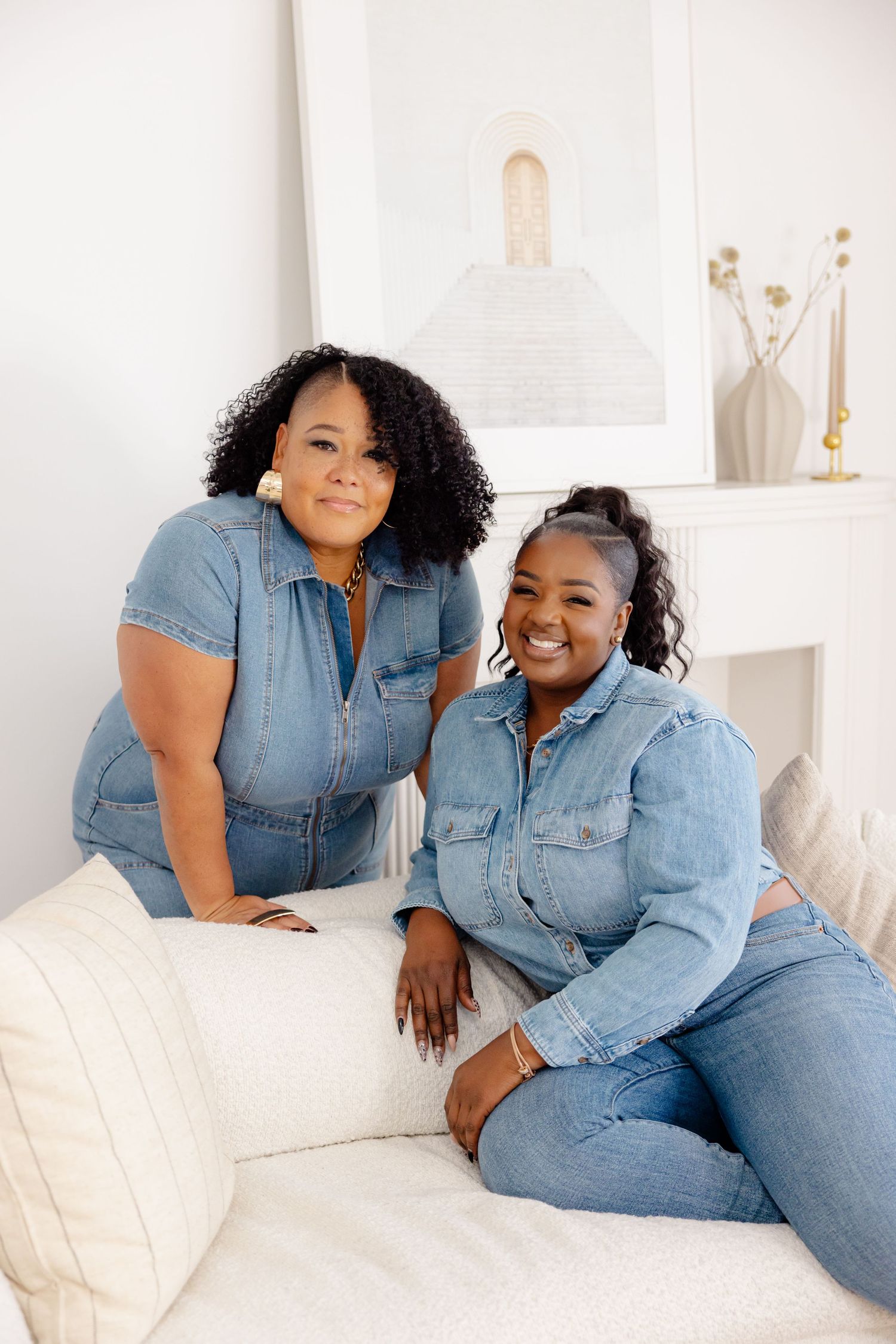 Two women wearing matching denim outfits pose while sitting on a white couch in a minimalist room.