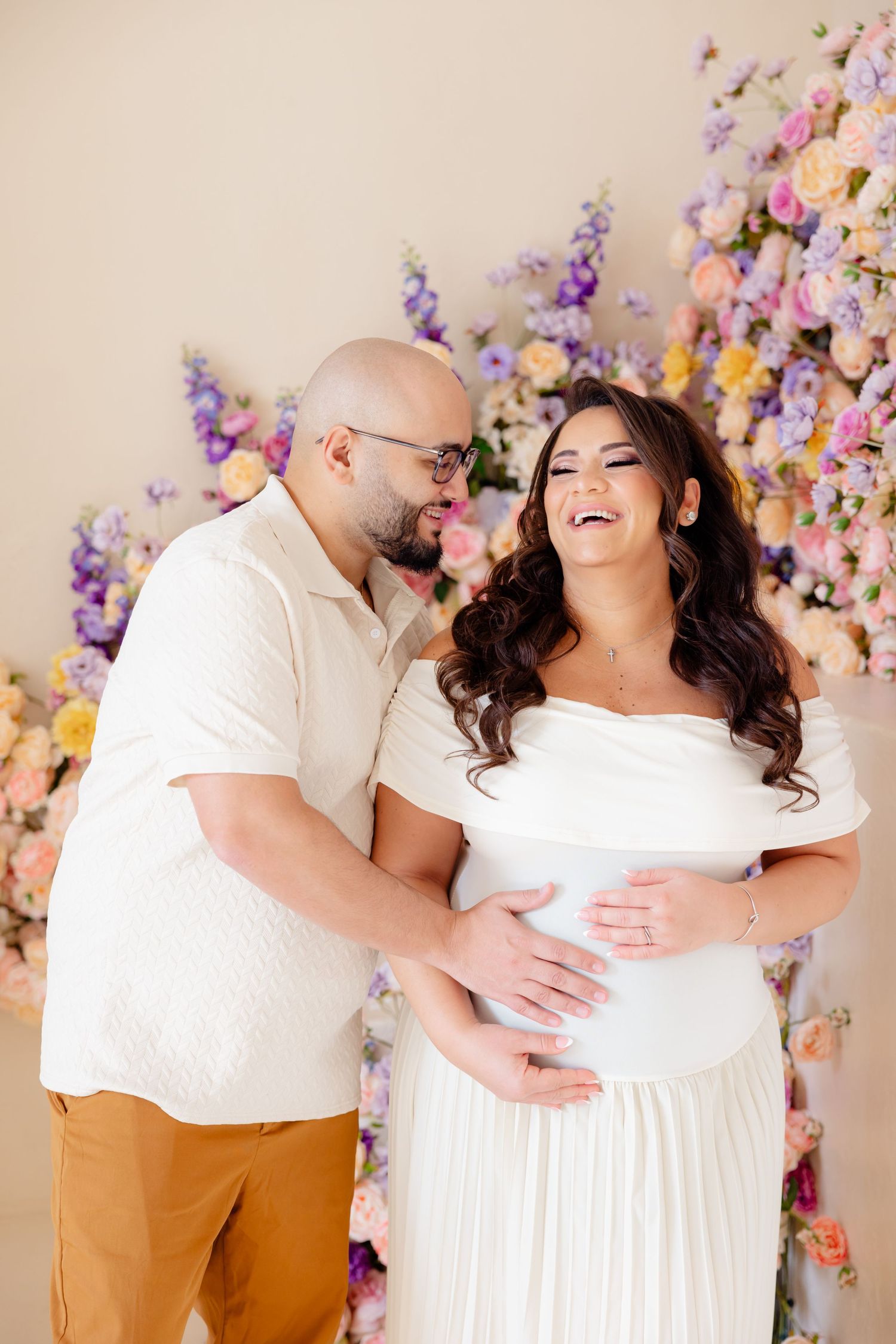 A joyful maternity photo featuring a couple in white outfits laughing together against a floral backdrop.