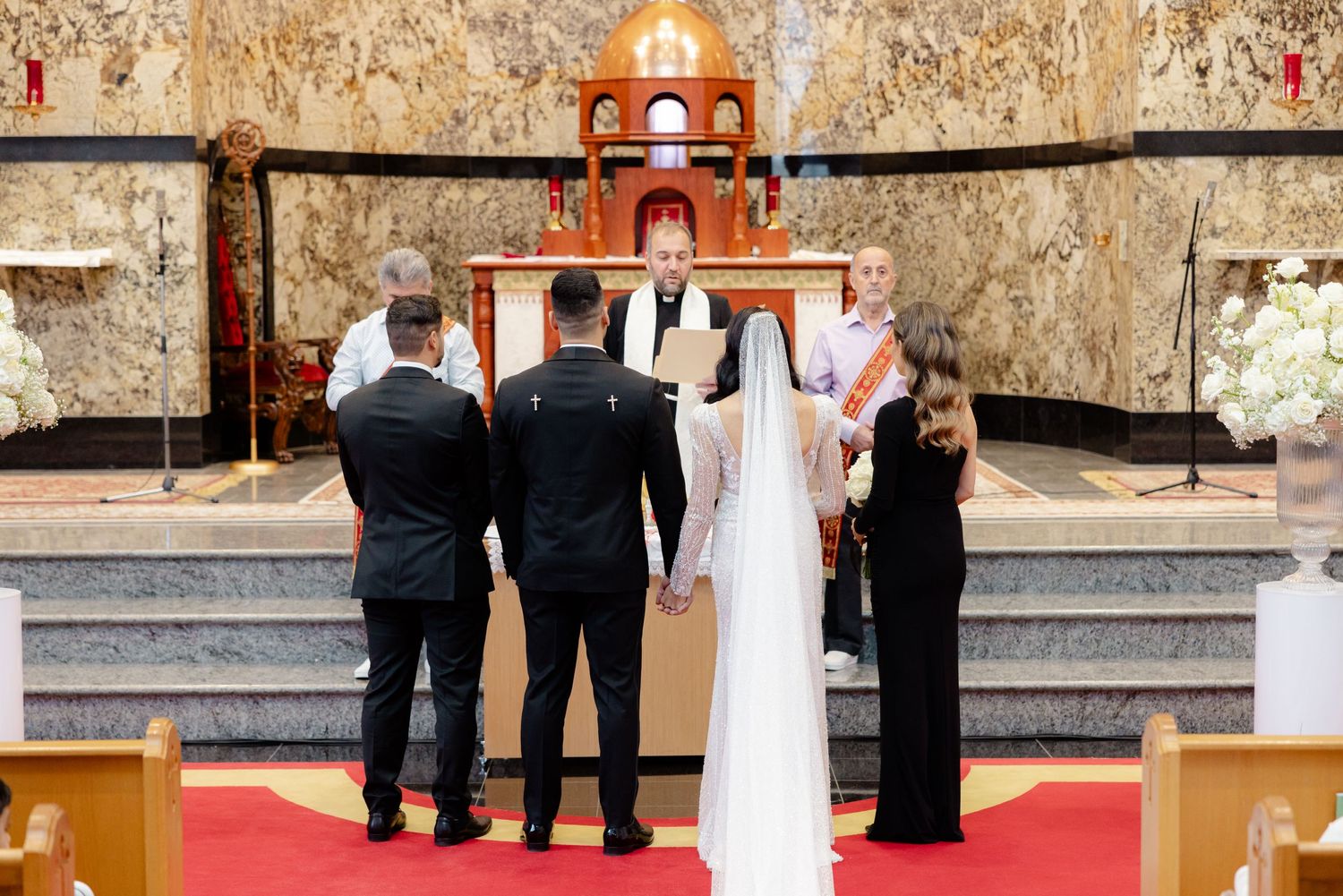 Wedding party stands at church altar during ceremony with gold-toned walls and red carpet.