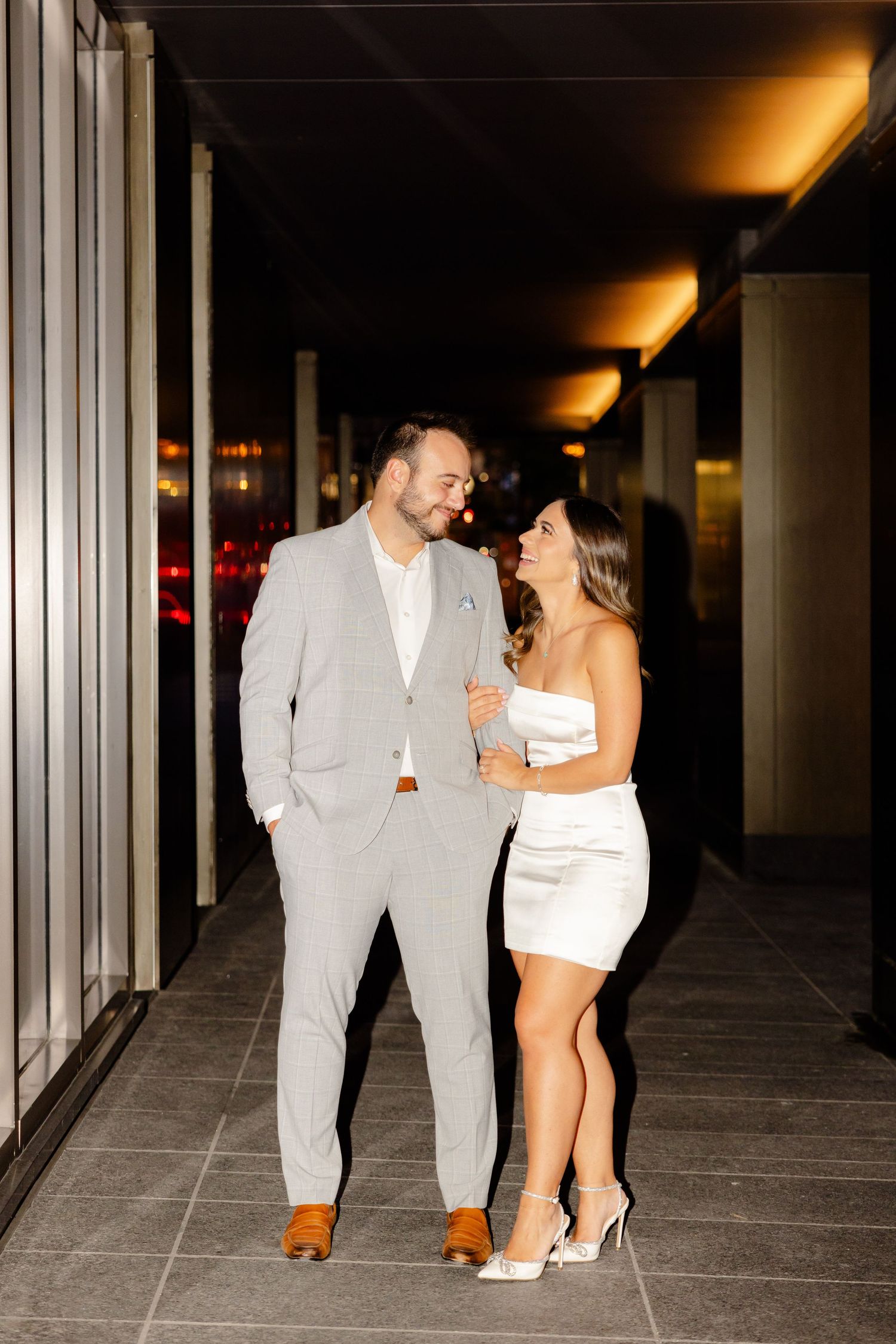 Happy couple in formal attire sharing a laugh in a modern hallway setting.
