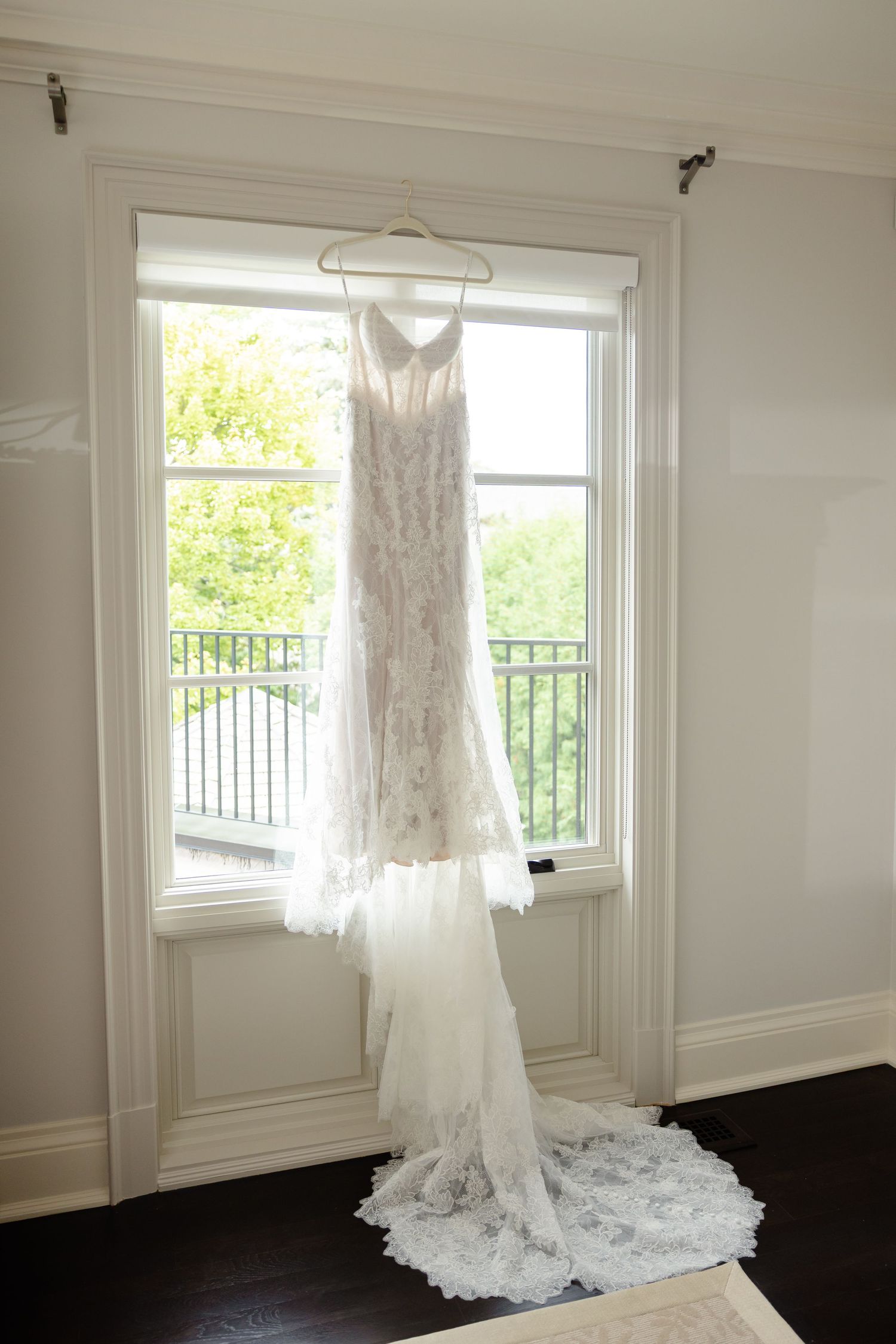 A white lace wedding dress hangs elegantly by a bright window with natural light streaming in.