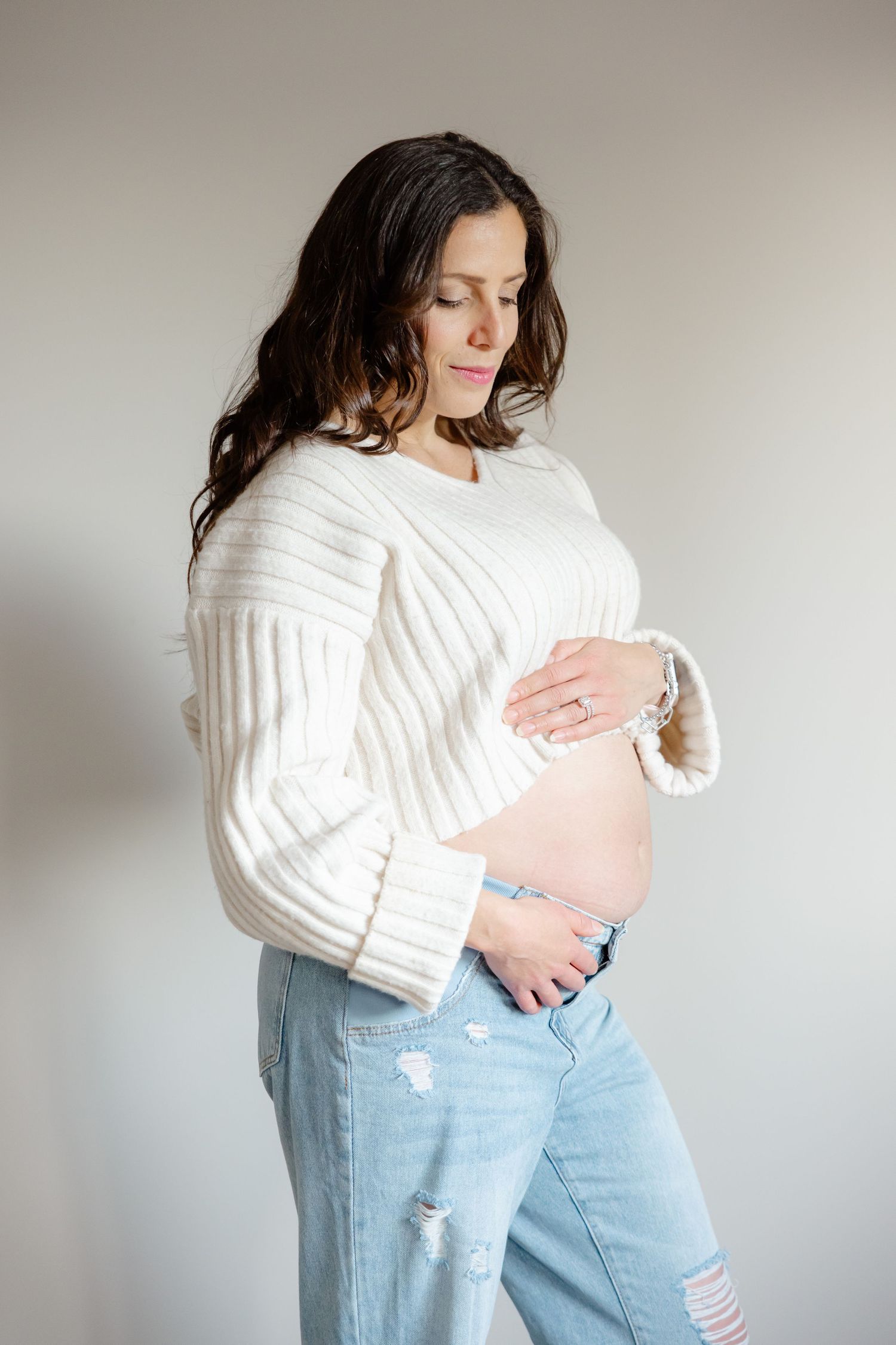 Pregnant woman in white sweater and jeans cradling her baby bump against a light background.