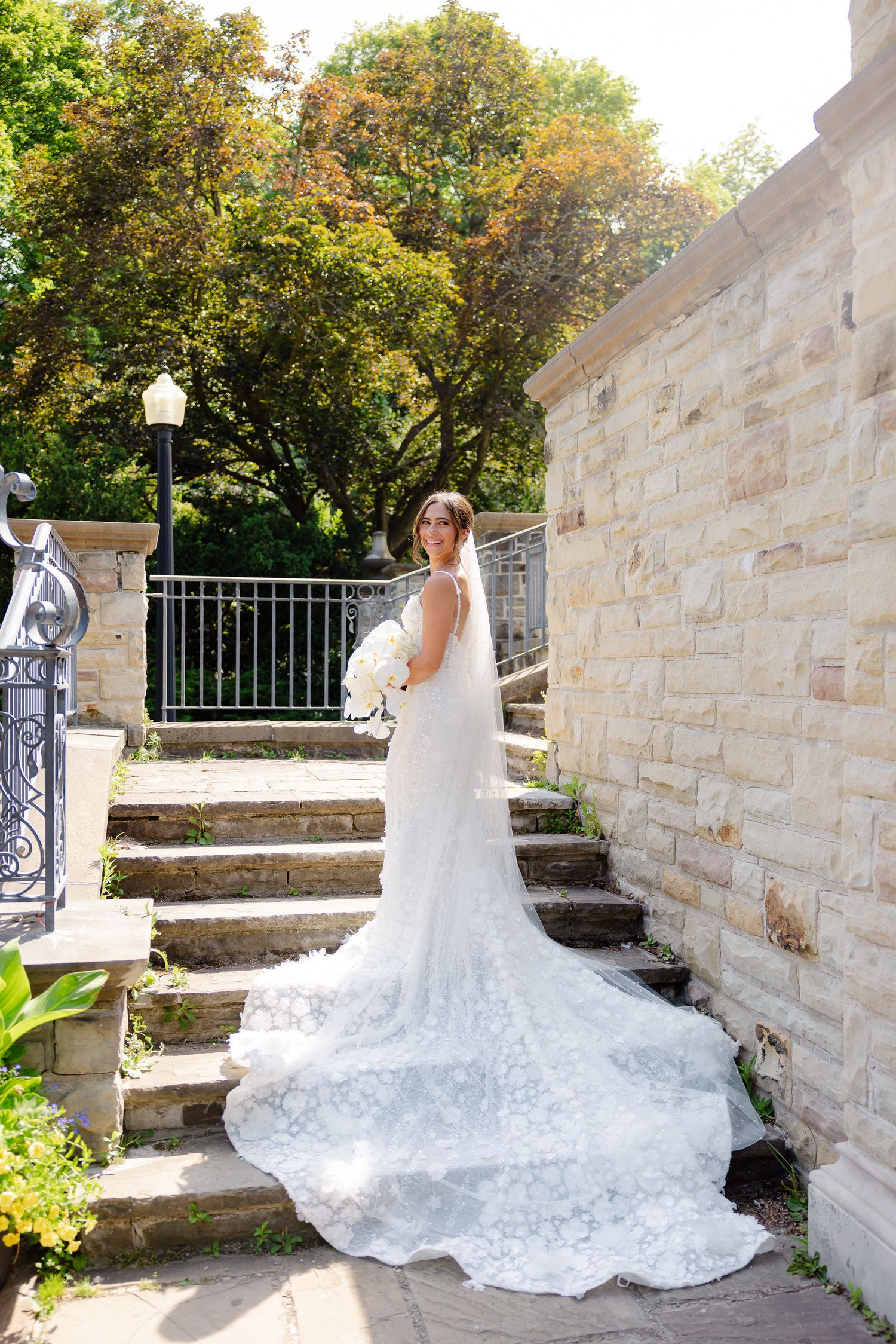 Elegant white lace wedding dress with long cathedral train cascading down stone steps in an outdoor garden setting.