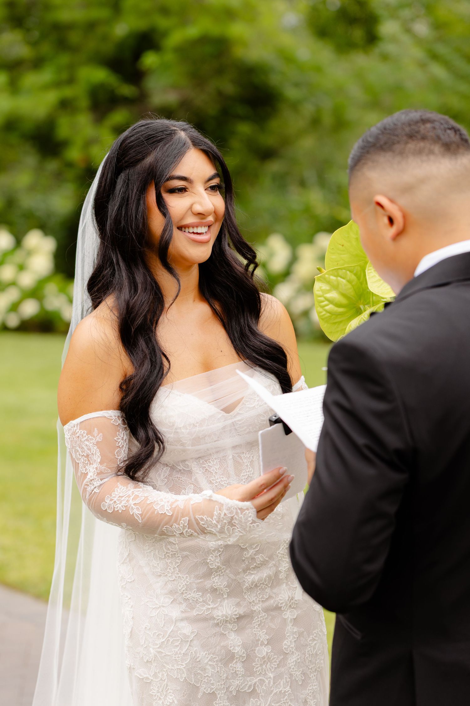 A bride with long dark wavy hair in a lace wedding gown exchanges vows during an outdoor ceremony.