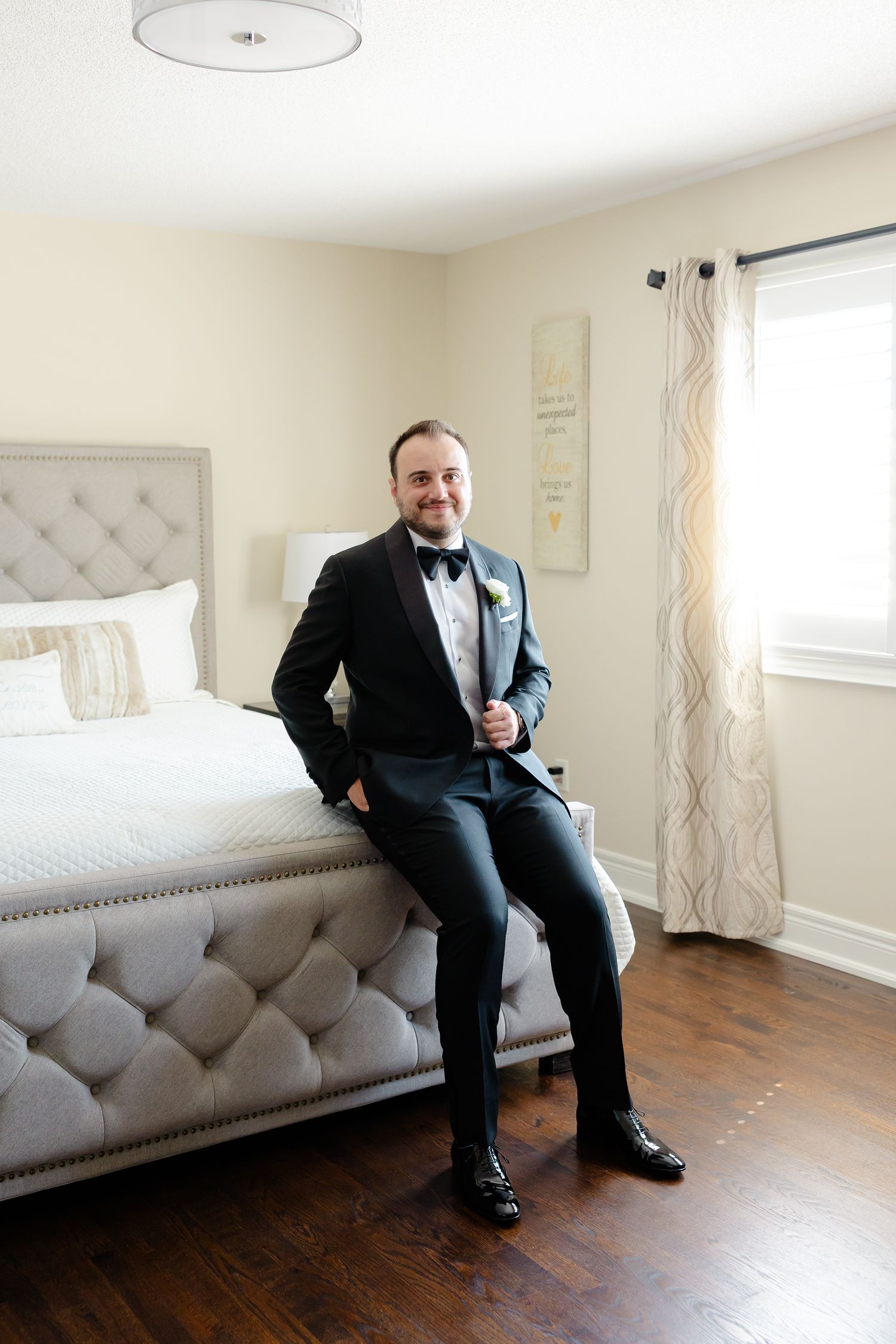 A groom in a black tuxedo and bowtie sits on the edge of a tufted grey bed in a bright bedroom with hardwood floors.
