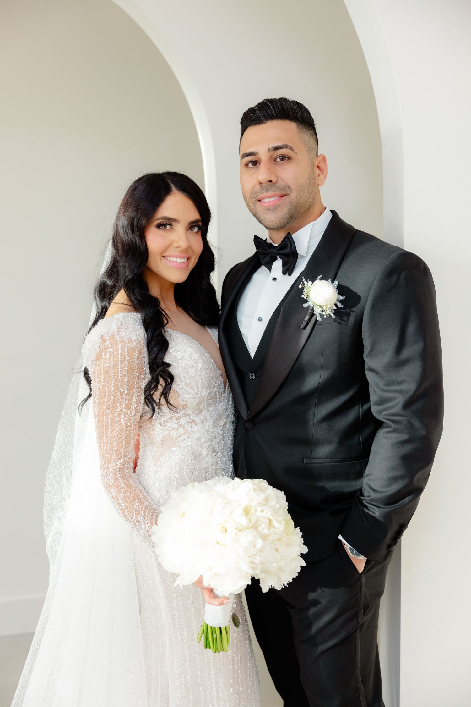 Wedding portrait of couple under white arch featuring elegant bridal gown and classic tuxedo.